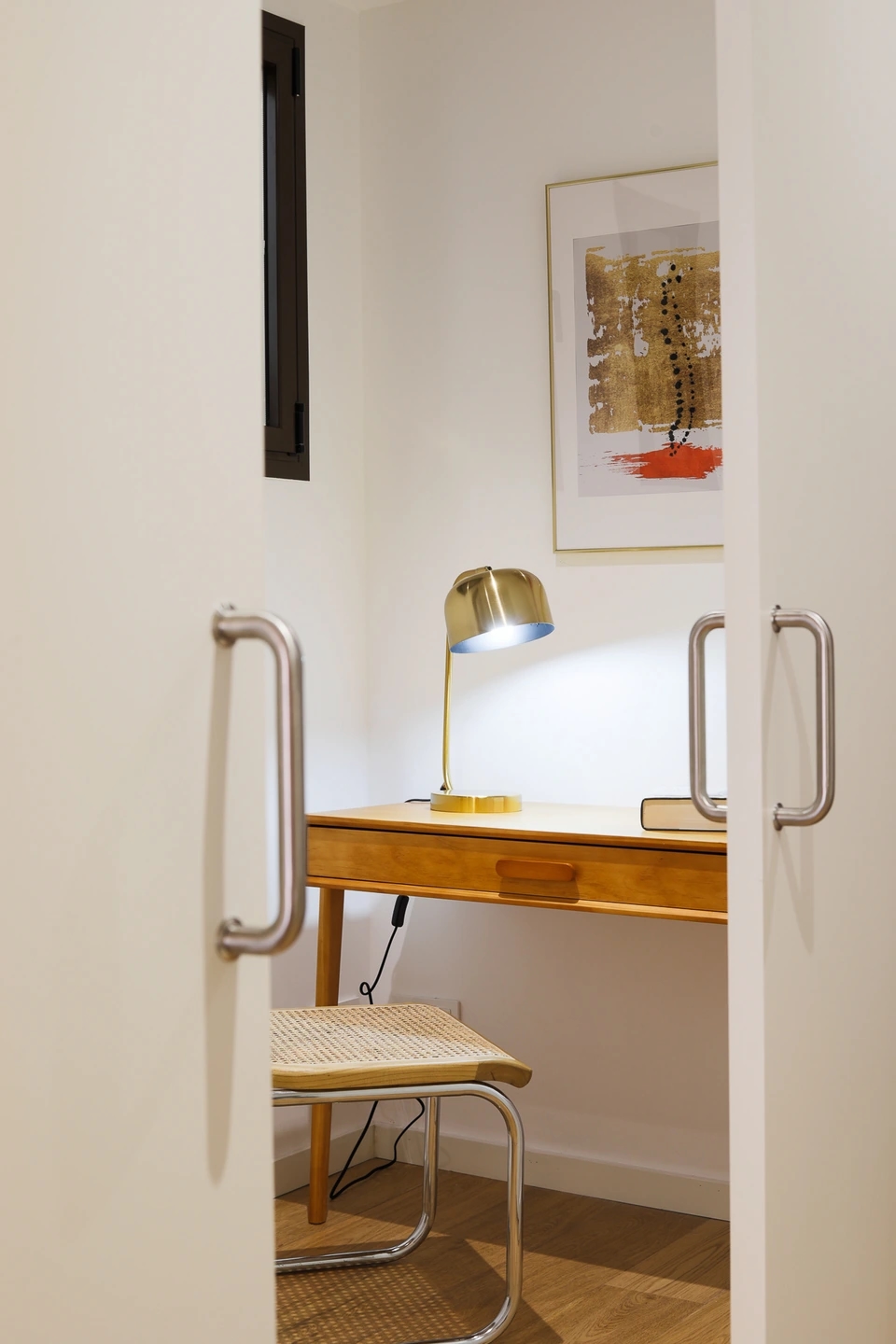 Fotografía de un escritorio de madera con lámpara dorada y silla de fibras naturales en un espacio de trabajo de Atelier Apartments.
