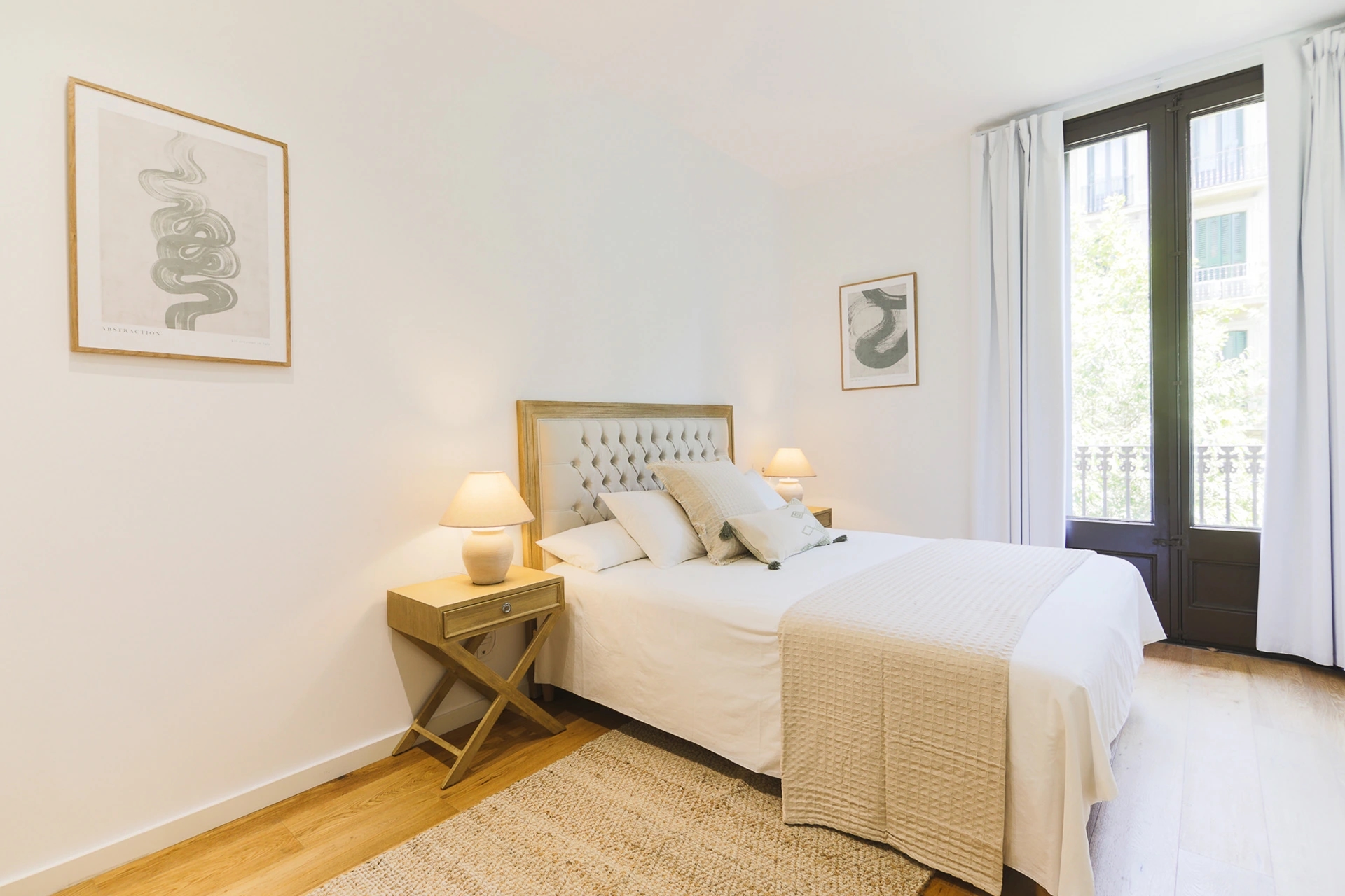 Fotografía de dormitorio luminoso en un apartamento de Atelier Apartments, con cama de cabecero tapizado, mesitas de madera, lámparas de cerámica y ventanal con cortinas blancas.