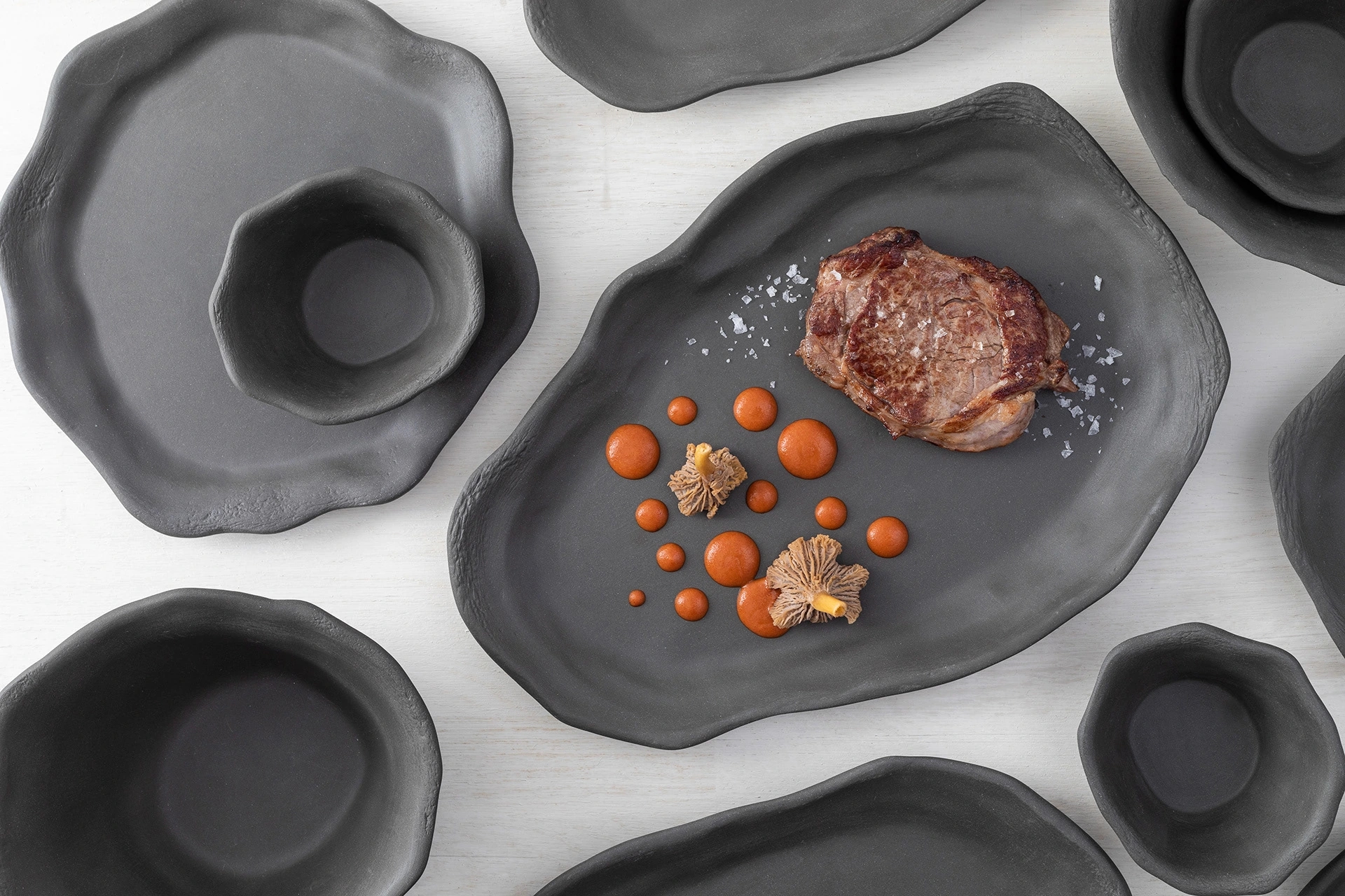 Composición con la vajilla negra "oyster" de oms y viñas. En el plato central hay un solomillo de ternera con un bosque de setas y salsa romesco.