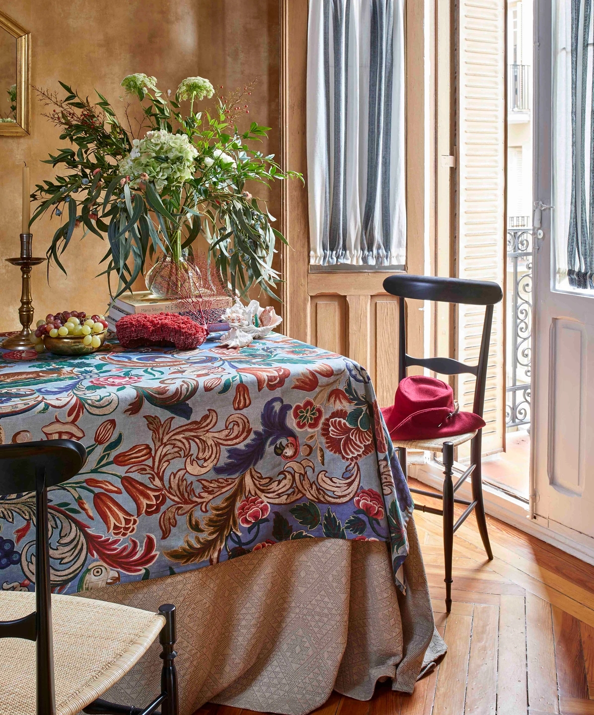 Fotografía de los tejidos de la colección Bloomsbury Collection - Güell Lamadrid y Melián Randolph. Mesa estilo medieval con sillas y sombrero de terciopelo con plumas de faisán, con jarro de eucaliptos y hibiscos en el centro de la mesa.