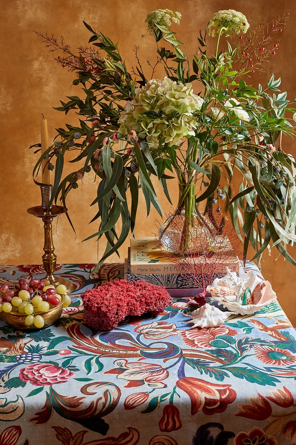Fotografía de los tejidos de la colección Bloomsbury Collection - Güell Lamadrid y Melián Randolph. Mesa estilo medieval con sillas y sombrero de terciopelo con plumas de faisán, con jarro de eucaliptos y hibiscos en el centro de la mesa.