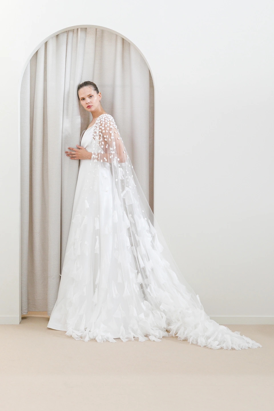 Fotografía de un vestido de novia de la colección Peter Langner con capa de tul bordada con flores en relieve, retrato largo en fondo neutro con un arco.