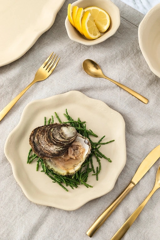 Mesa parada con cubertería de oro y la vajilla oyster color arena con unas almejas en el plato.