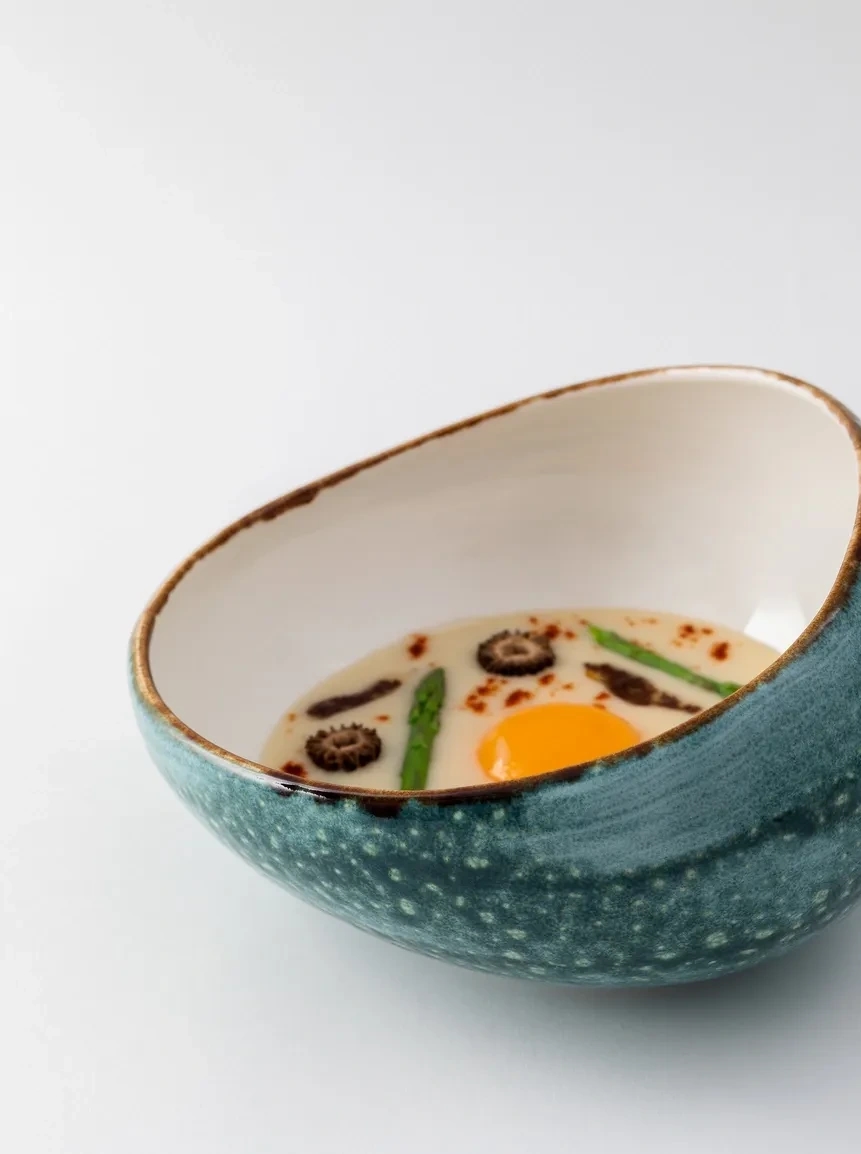 Composición con un bol en el centro y un plato de alta cocina que parece como un consomé con huevo, espárragos y setas. Chef Martin Lippo
