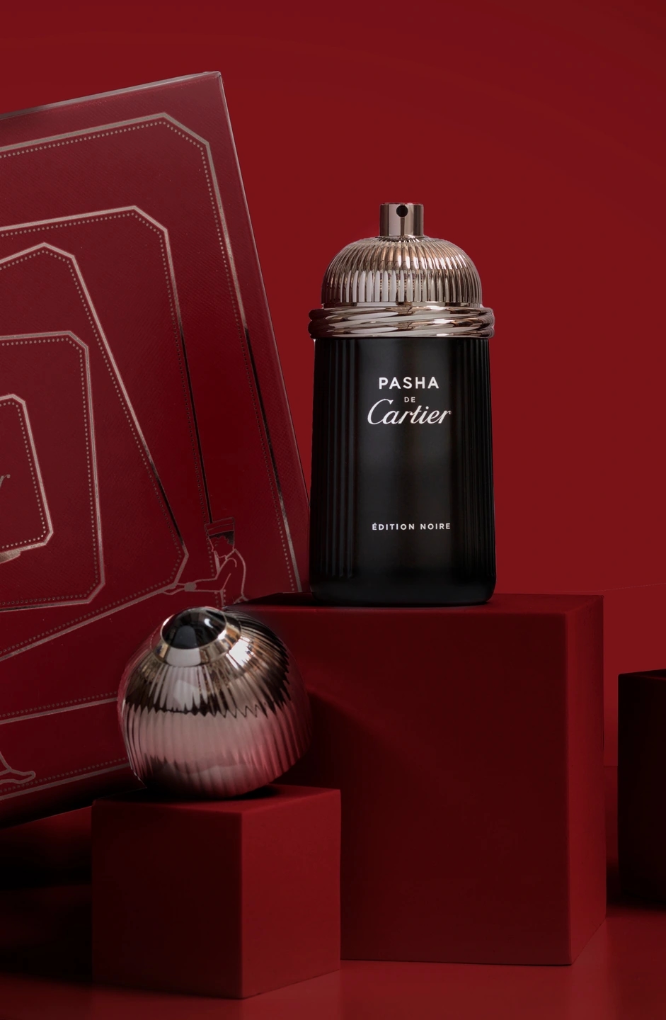 Composición oscura del perfume Cartier PASHA y la caja en la que viene envuelto. Fondo rojo oscuro. Fotografía para la marca de perfumerías Éclair.