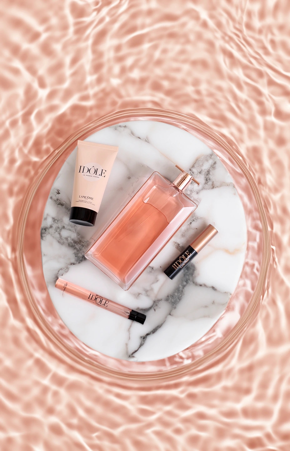 Fotografía zenital de perfume Idôle de Lancôme, crema de manos, perfume de viaje y máscara de pestañas. Encima de un mármol redondo y flotando en agua de color salmón.