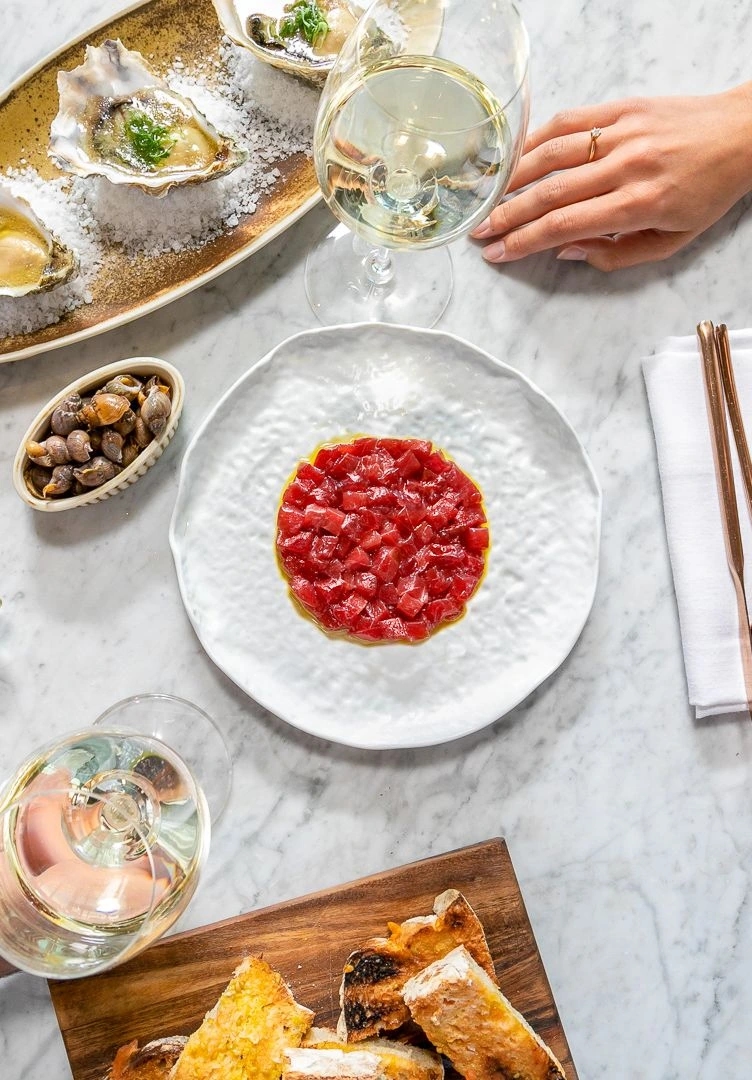 Mesa vista cenital con ostras, tartar de atún, encurtidos y copas de vino blanco sobre mármol.