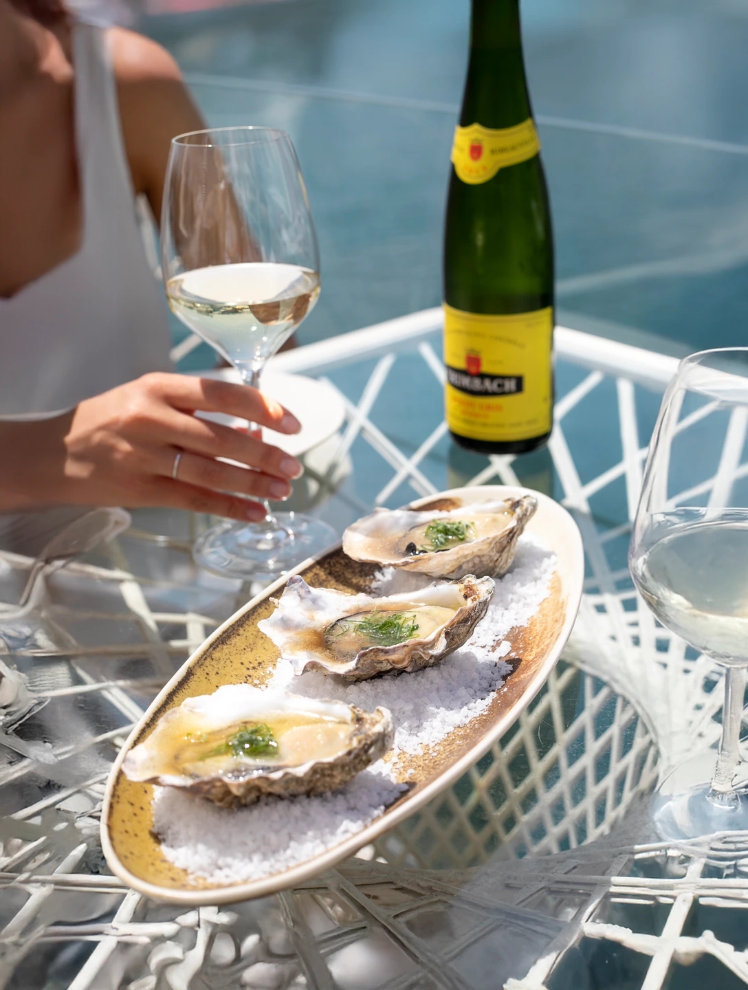 Comensal brindando con vino blanco junto a un plato de ostras sobre hielo en una mesa exterior frente al mar.