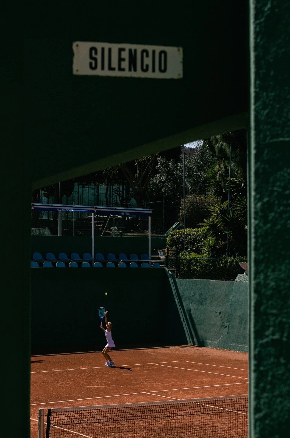 adolescente jugando a tenis en una pista profesional de tierra batida con equipación de la marca ROGGIO de color blanco . Hay un cartel que pone SILENCIO