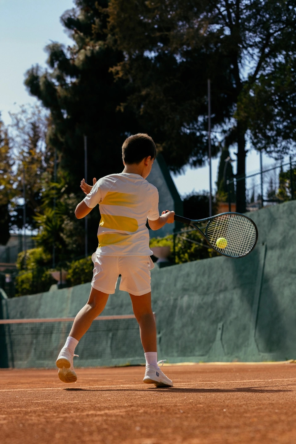 niño jugando a tenis en una pista profesional de tierra batida con equipación de la marca ROGGIO de color blanco y amarillo