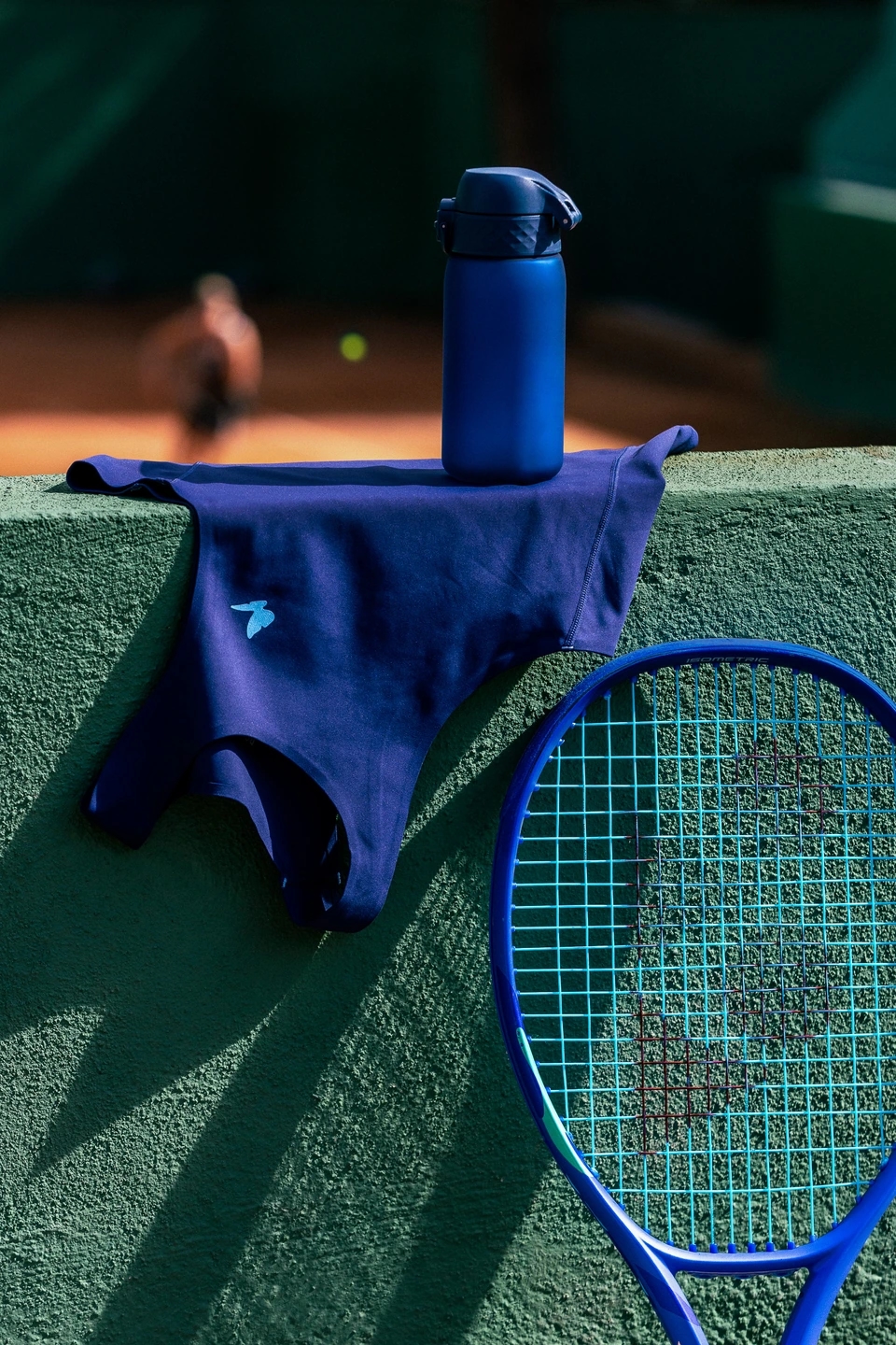Camiseta azul eléctrico Roggio apoyada en un muro verde abeto con una raqueta y una botella de agua del mismo color. De fondo un jugador de tenis golpea la pelota.
