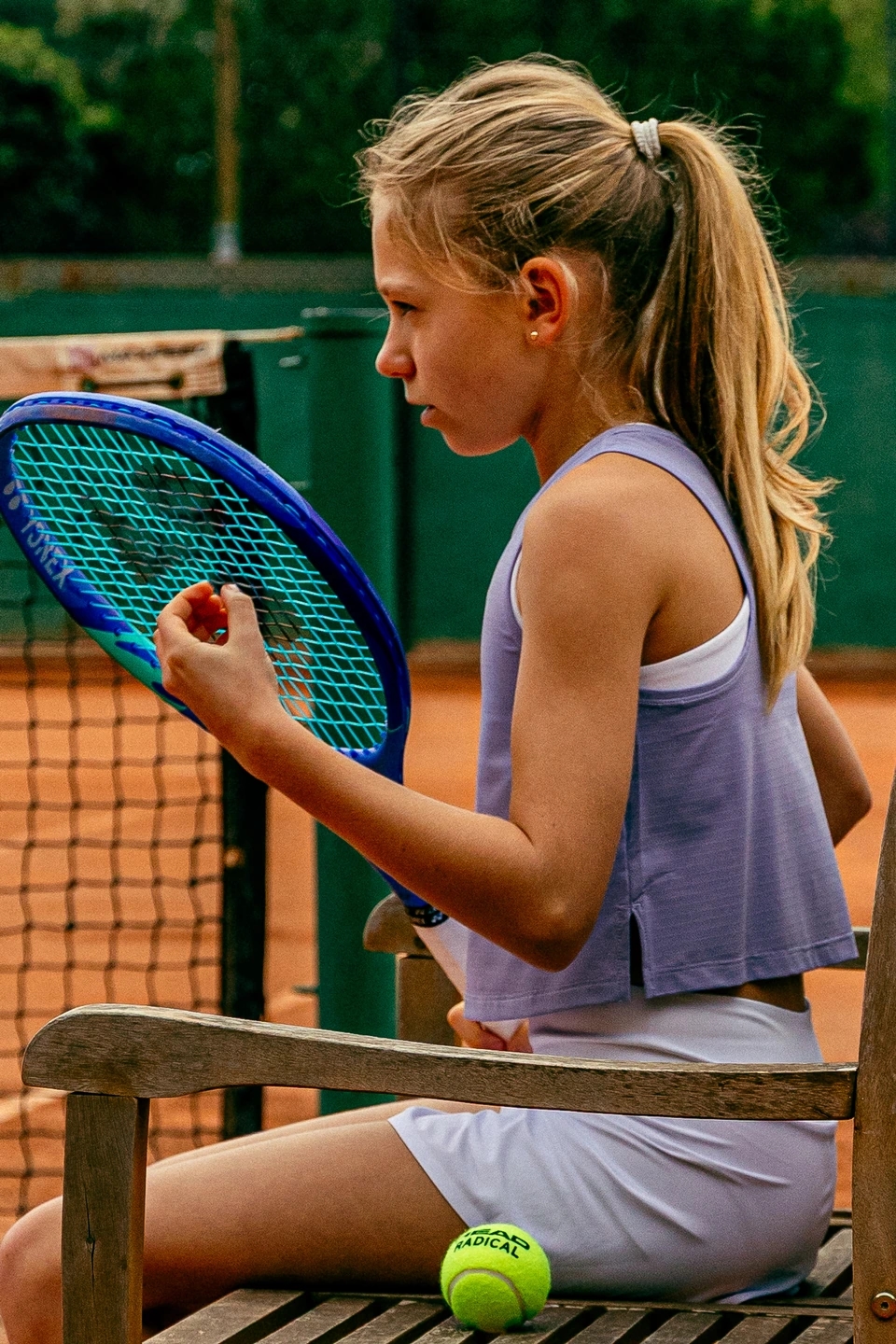 adolescente sentada preparándose para jugar a tenis en una pista profesional de tierra batida con equipación de la marca ROGGIO de color blanco y lila 