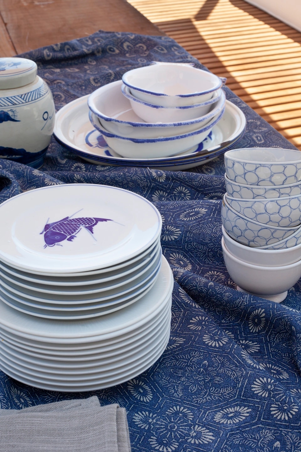 Mantel de la Colección La Cala de Güell Lamadrid con platos apilados encima y una carpa azul dibujada en el último plato. Estética veraniega mediterránea.