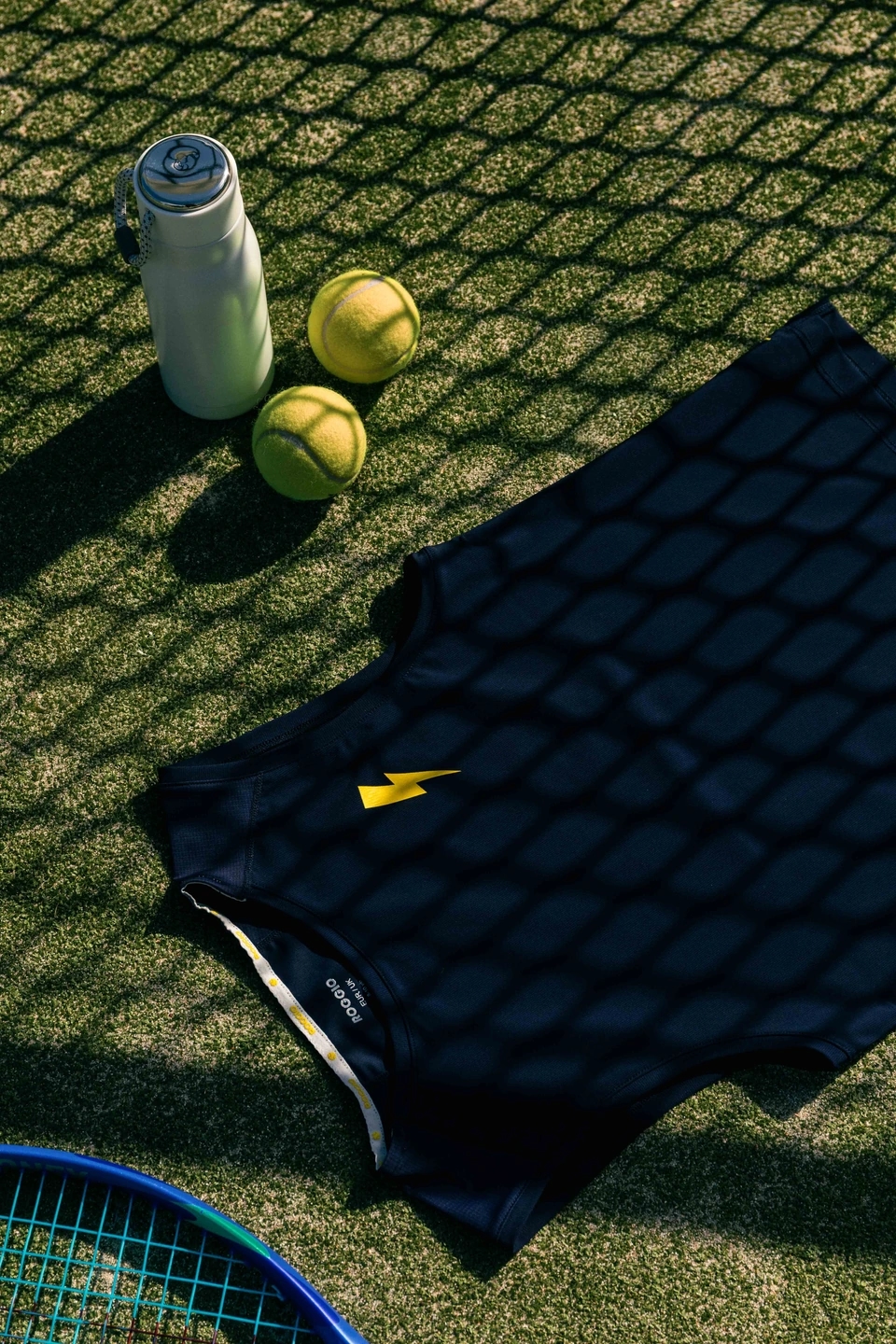 Camiseta azul oscuro Roggio en un campo de hierba de tenis con dos pelotas y una botella blanca. 