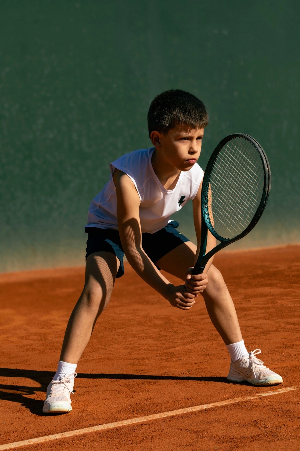 Niño con ropa Roggio en posición de defensa con una raqueta de tenis esperando el saque en una pista de tenis de tierra batida