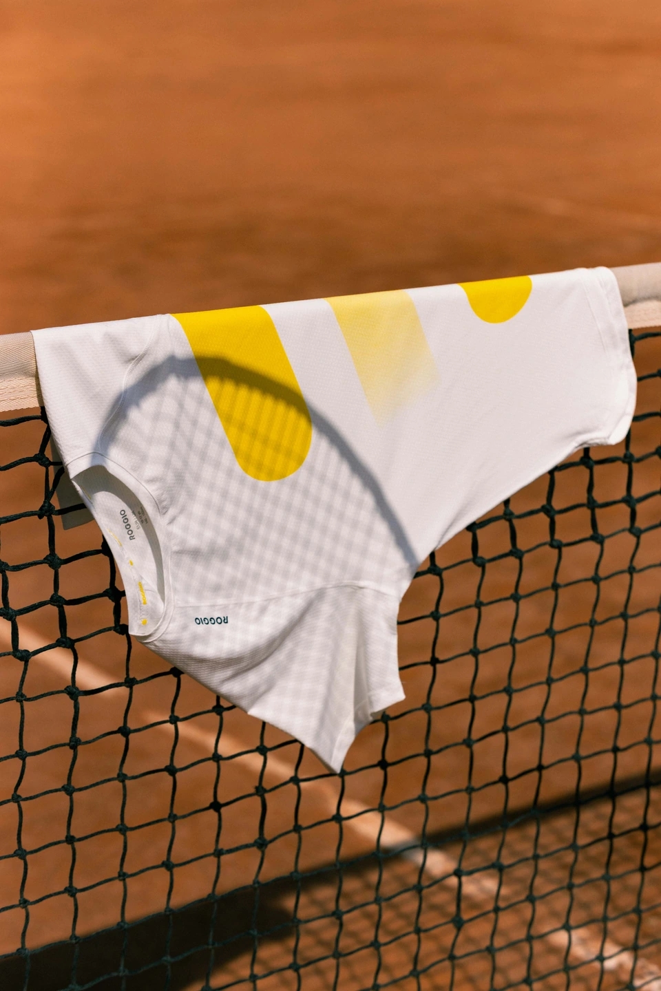 camiseta de la marca Roggio colgando en una red de tenis, en una pista de tierra batida. Se marca la sombra de una raqueta en la camiseta