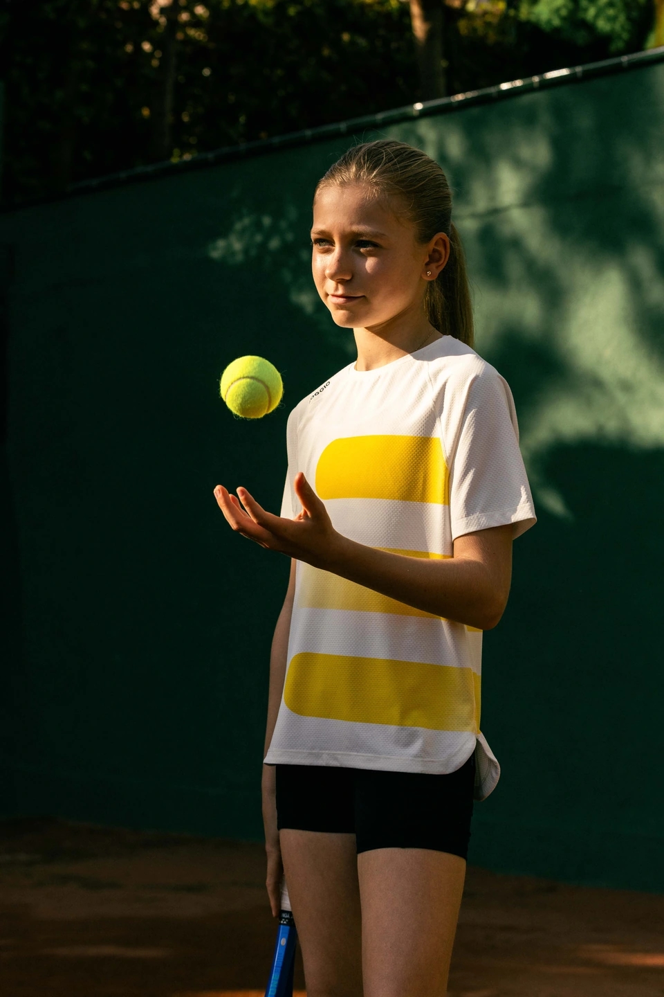 Detalle de una pelota de tenis heat con una raqueta de tenis y una niña sujetando la pelota. Va vestida con ropa roggio.