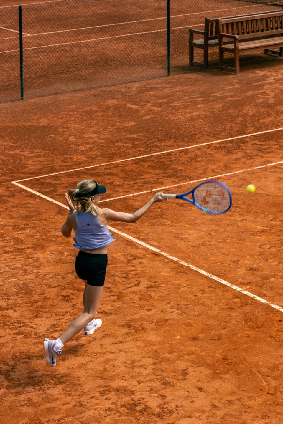 Adolescente tenista profesional en una pista de tenis de tierra batida compitiendo. Va vestida con ropa de la marca Roggio de color lila