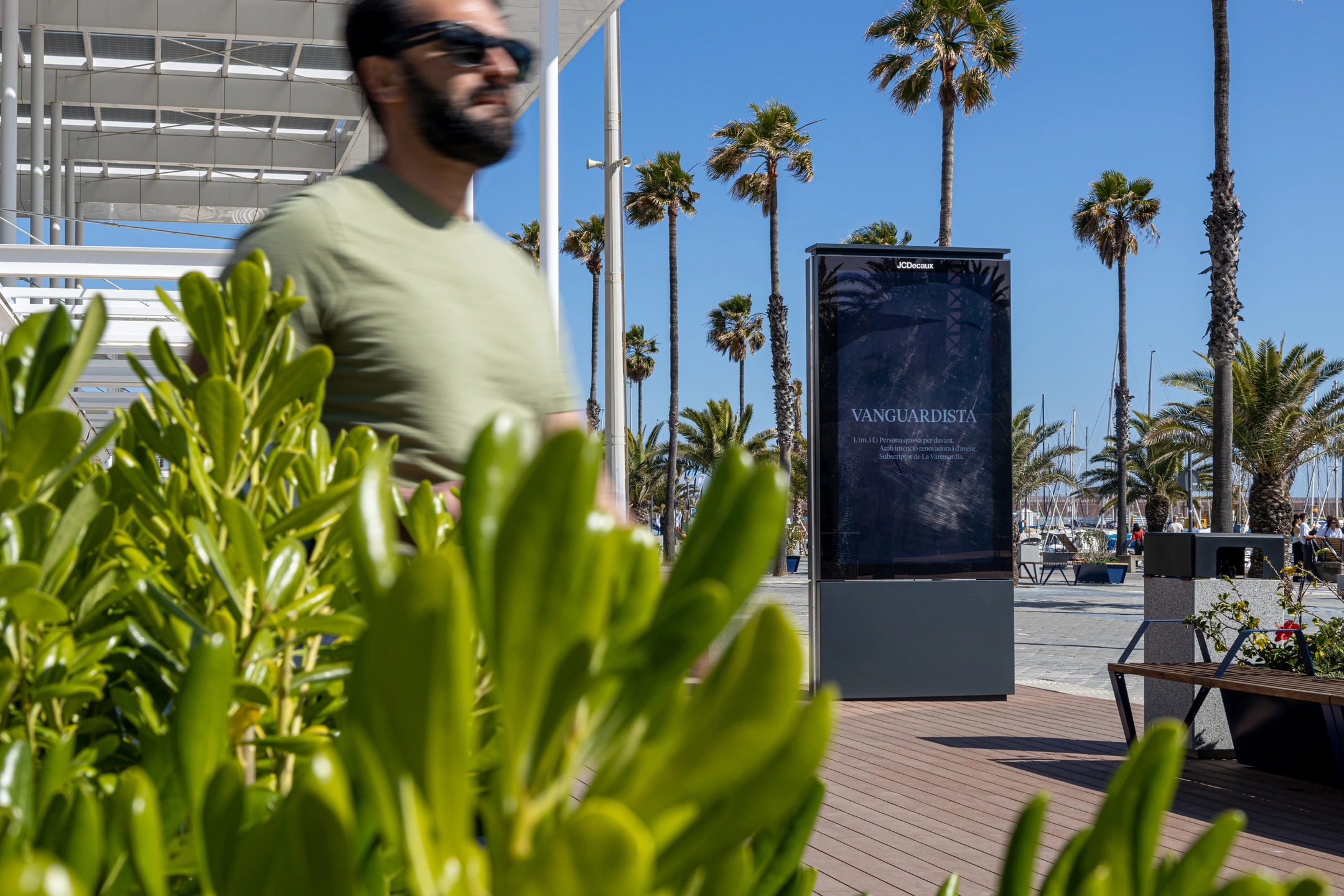 Cartel publicitario en el Port Olímpic de Barcelona con palmeras y cielo despejado.