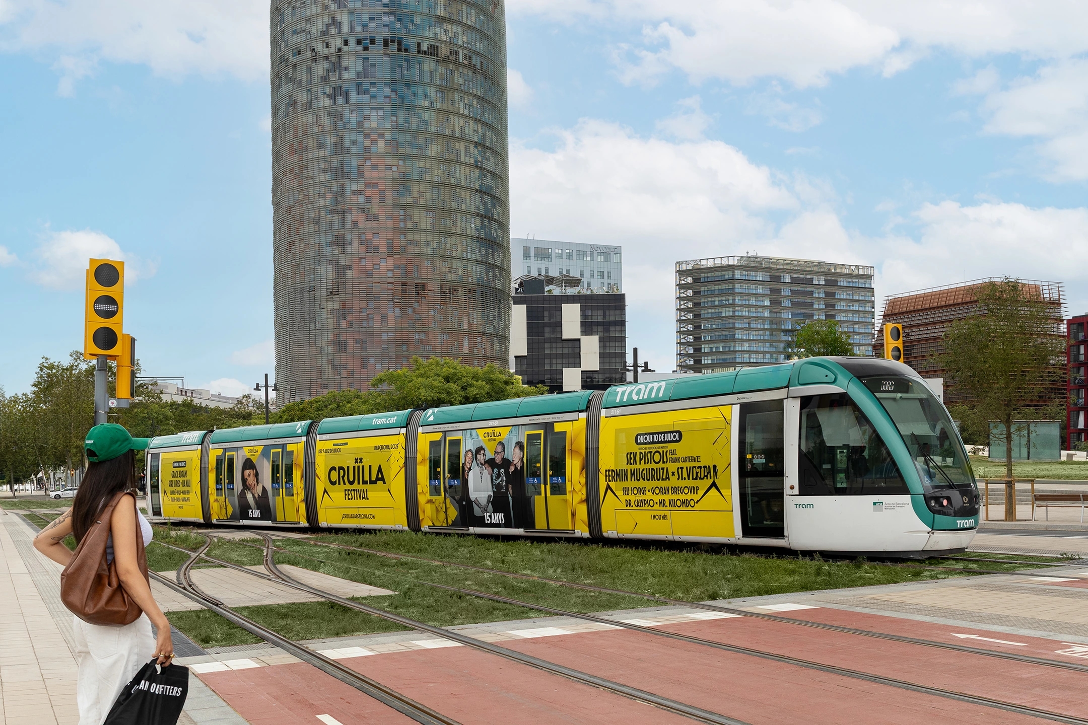 Tranvía cruzando Barcelona en la plaza de las glorias con la torre de las aguas de barcelona de fondo. Mujer con gorra verde lo mira.