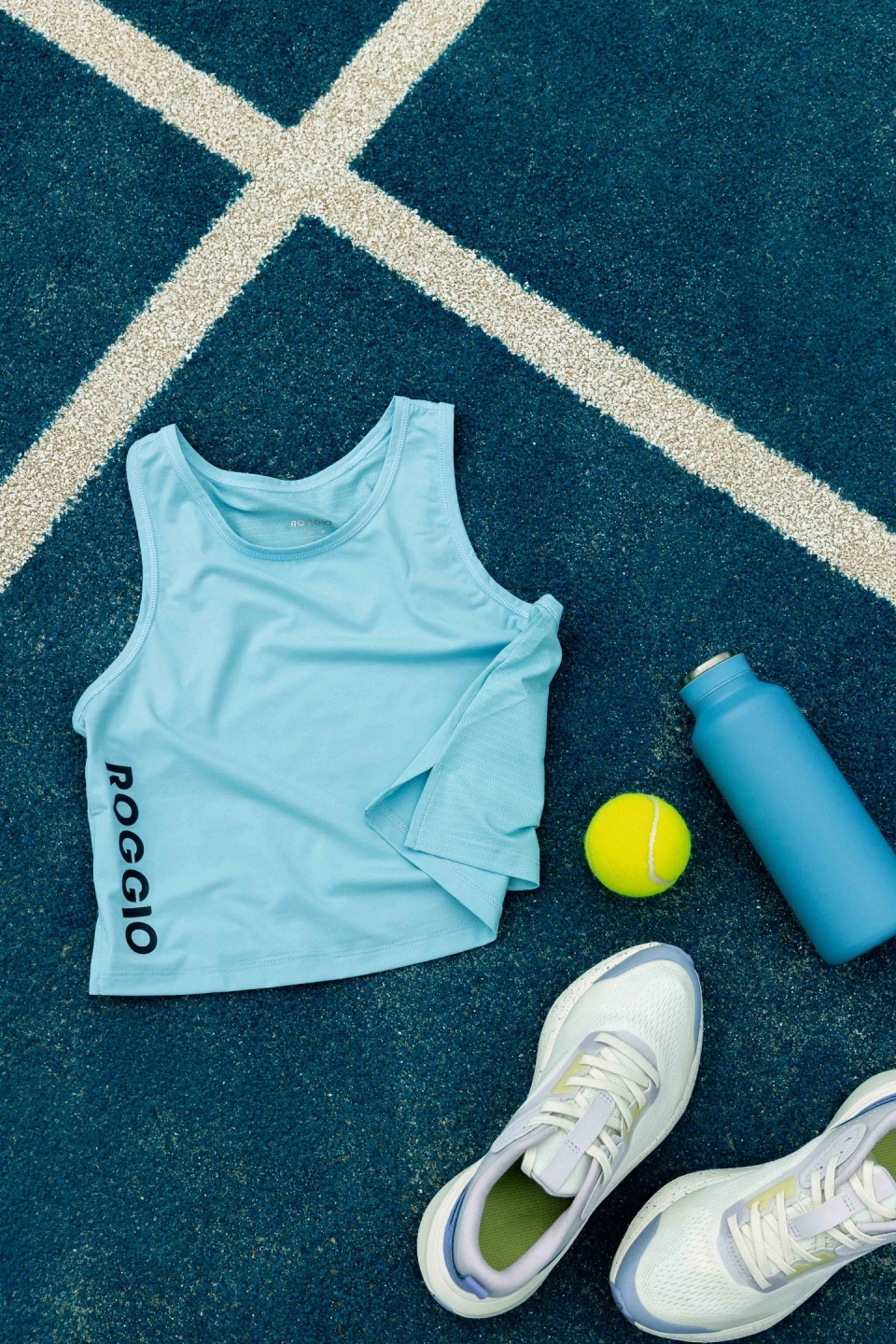 Bodegón de camiseta azul cielo Roggio, bambas decathlon blancas, pelota de tenis head y botella de metal azul en una pista de fronton. 