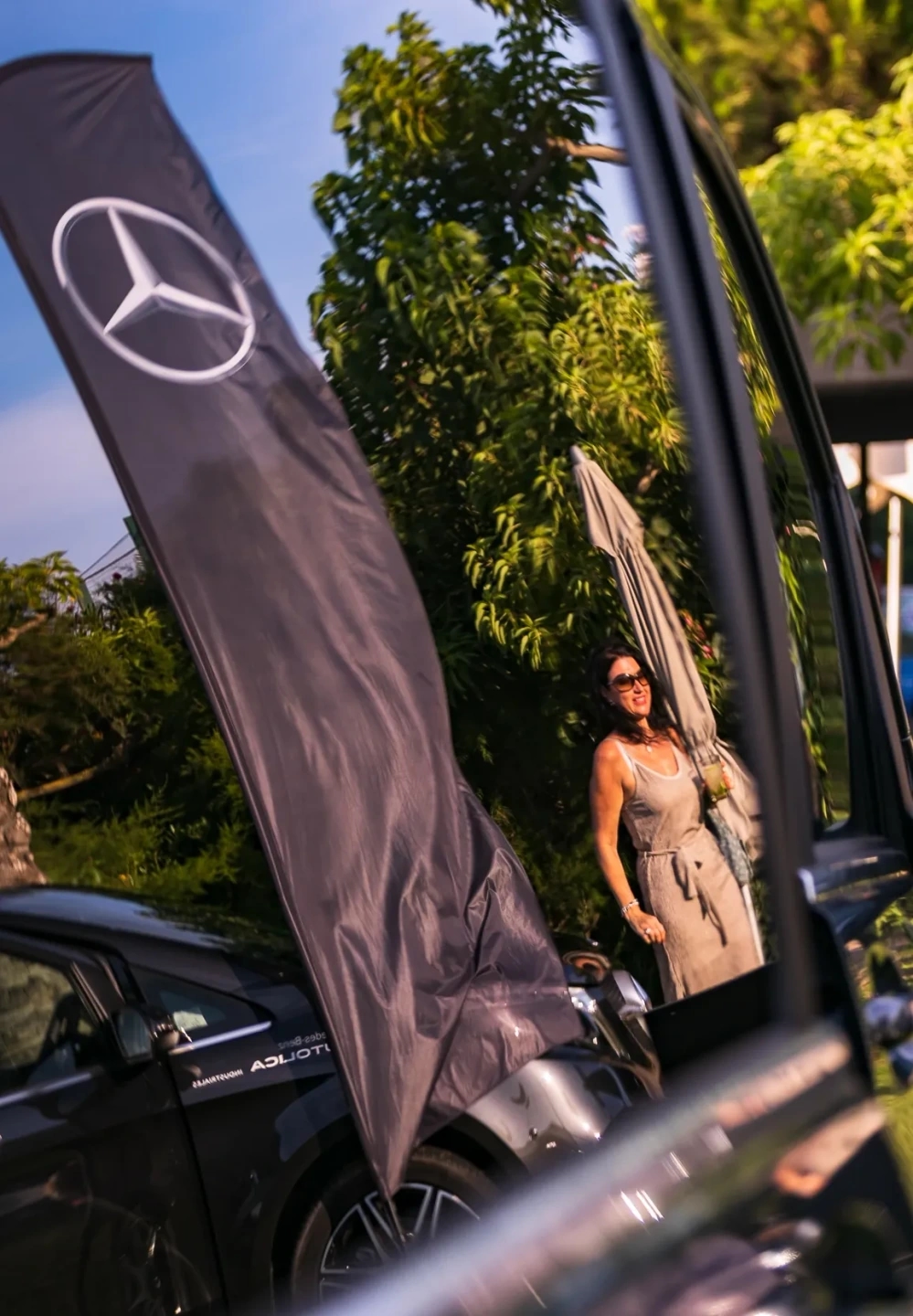 Reflejo de un vehículo Mercedes Autolica con bandera corporativa y una asistente del evento al fondo, en un entorno exterior ajardinado.