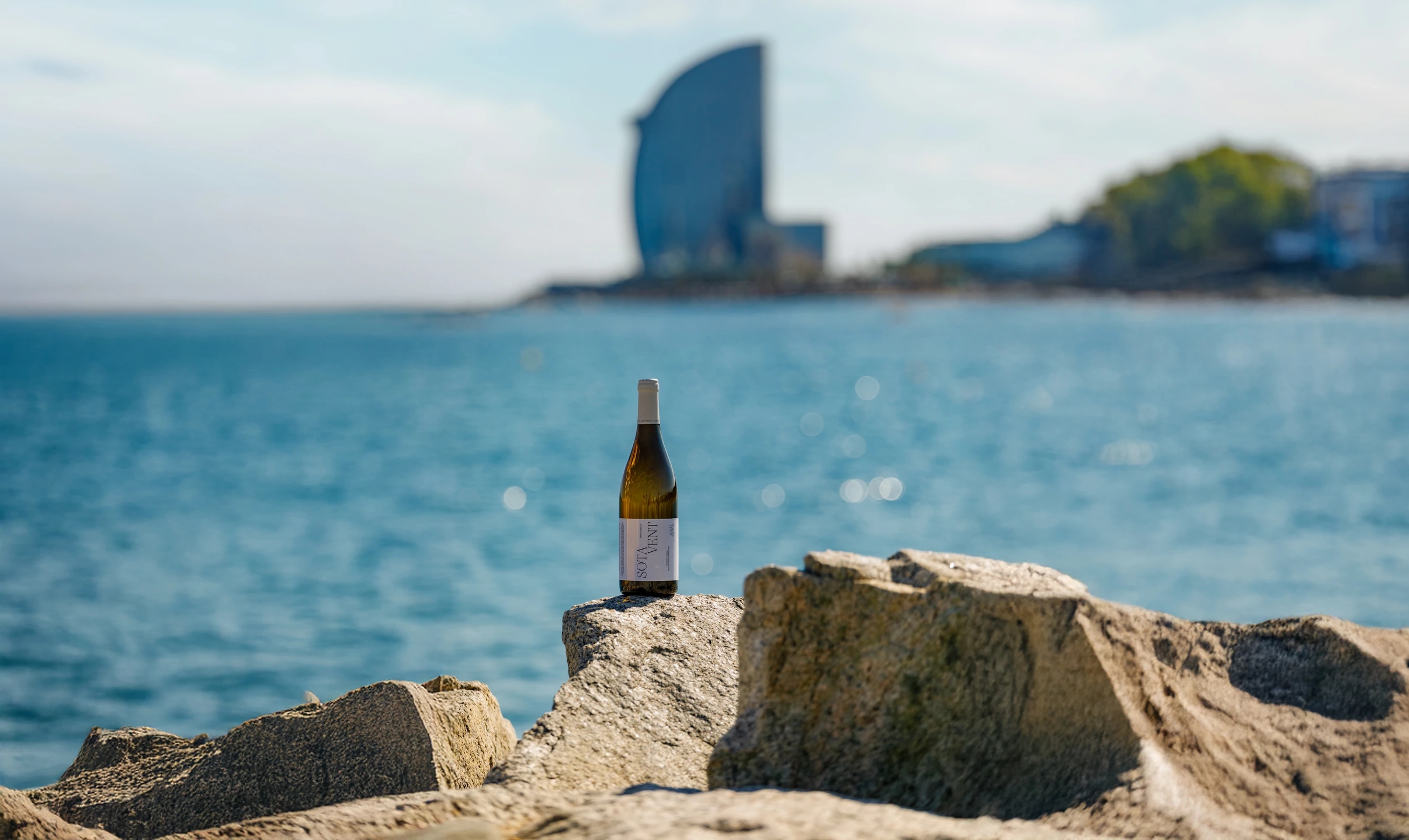 Botella de vino blanco Jaume sardà apoyado en unas rocas del mar mediterraneo. Hotel vela W de fondo