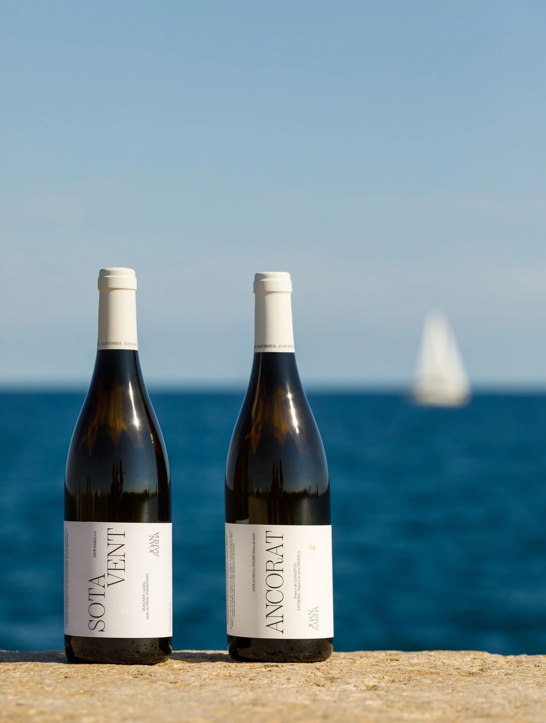Fotografia de los vinos Ancorat y Sotavent en un espigón de Barcelona, mar mediterráneo y unos barquitos desenfocados de fondo.