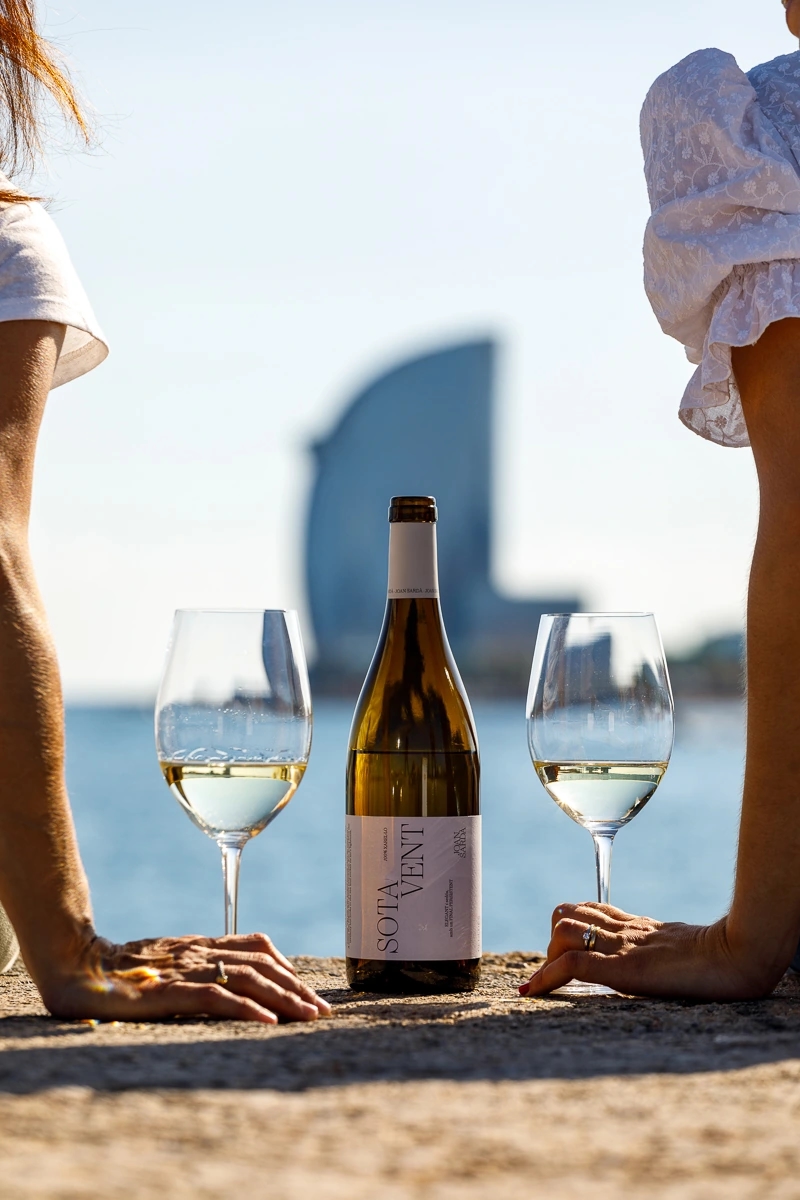 Pareja con dos copas de vino blanco "Sota vent" de la marca Joan Sardà y la botella de vino mientras ven el atardecer en un espigón de barcelona con vistas al hotel vela.
