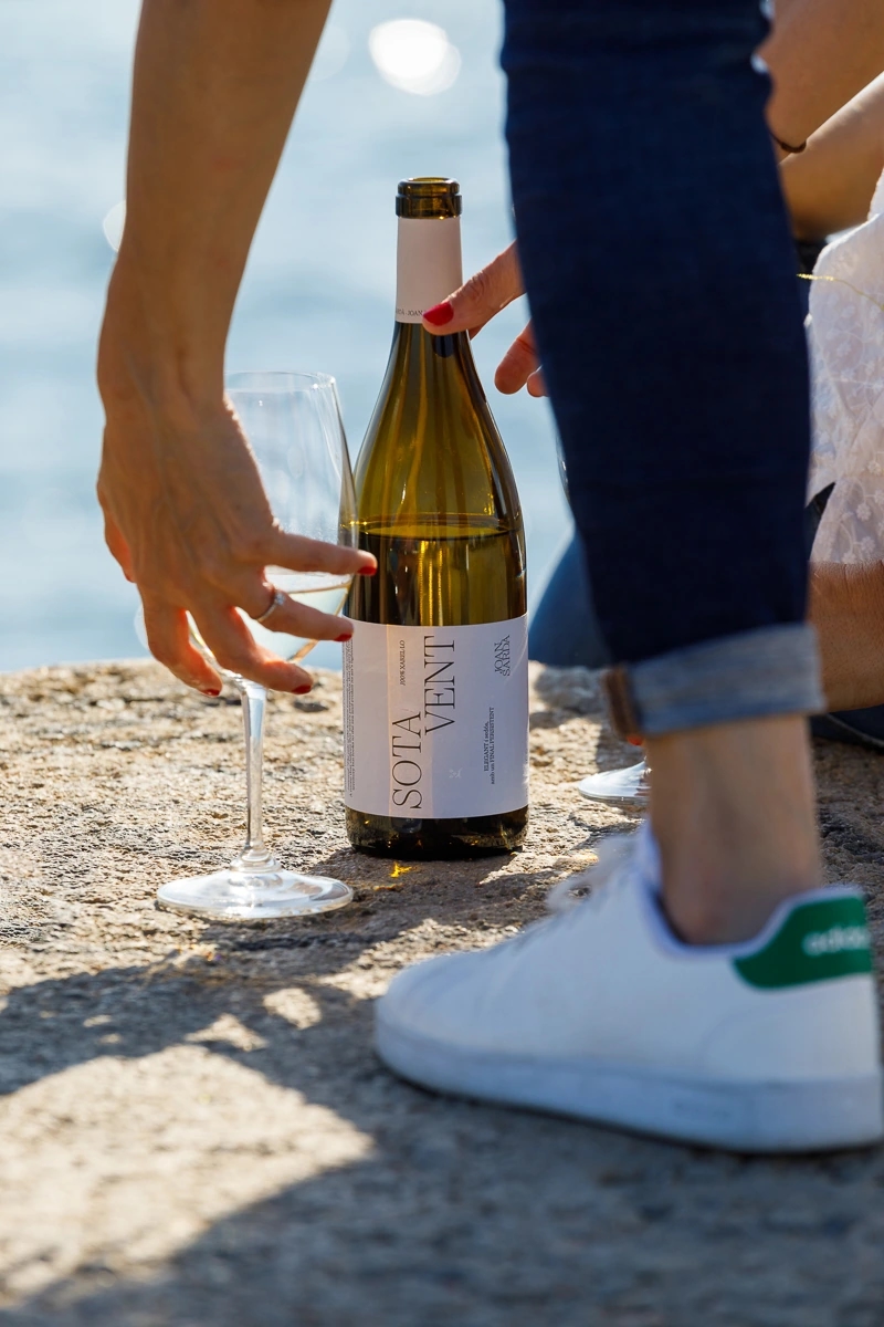 Mujer recoge dos copas de vino blanco "Sota vent" de la marca Joan Sardà y la botella de vino en un espigón de barcelona con vistas al mediterráneo.