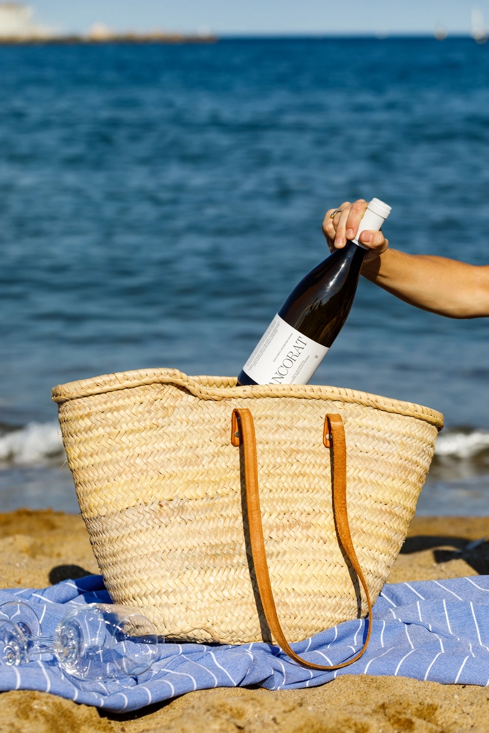 Mujer saca una botella de vino "ancorat" de Joan Sardà de un cesto de paja típico de playa. Está sentada en un pareo de playa delante del mar mediterráneo.