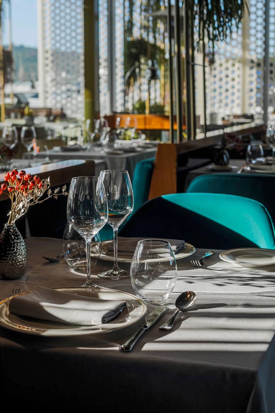 Detalle de mesa puesta con copas y servilletas en un restaurante con luz natural y sillas turquesa.