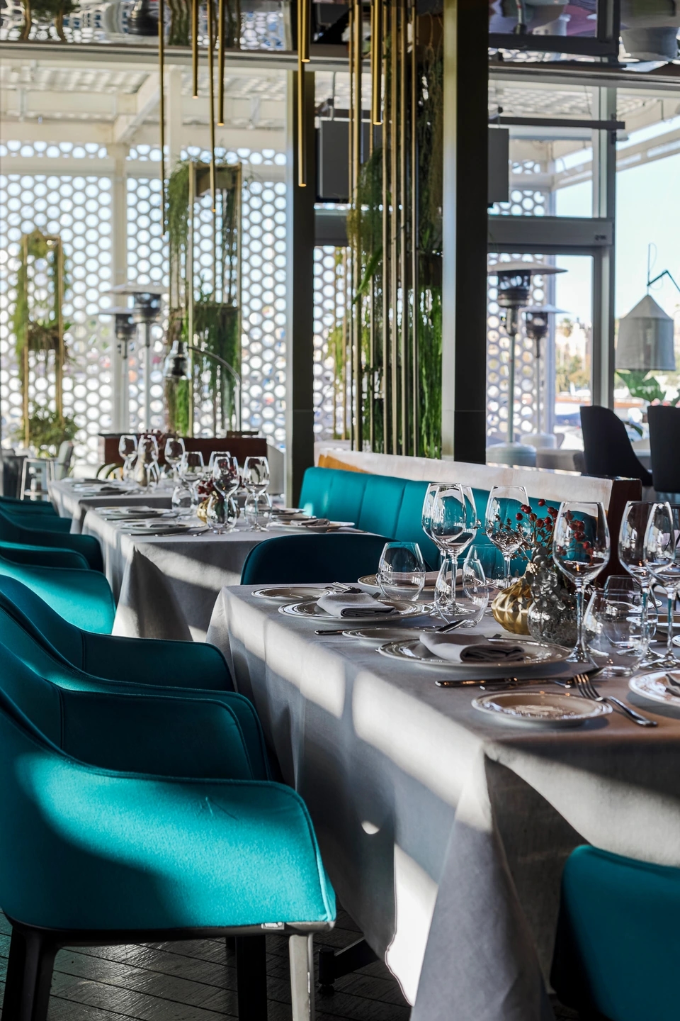 Mesa de restaurante Marina Port Vell con vajilla y copas, sillas turquesa y luz natural entrando por los ventanales.