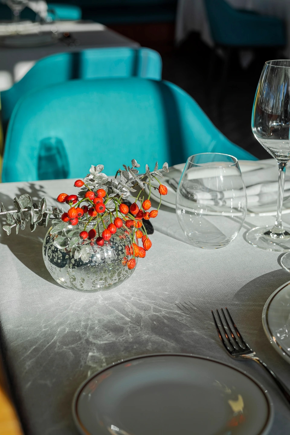 Centro de mesa con bayas rojas en jarrón de cristal sobre mantel gris en un restaurante luminoso.