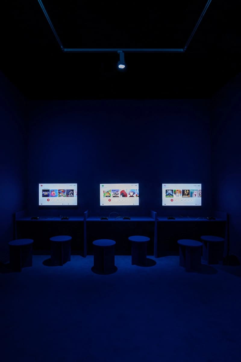 Fotografia de la escenografia de la exposición de Nueva pantalla, el videojuego en Cataluña. Fondo azul oscuro, escenografía con luz muy tenue y tres pantallas con 6 asientos para jugar a videojuegos.