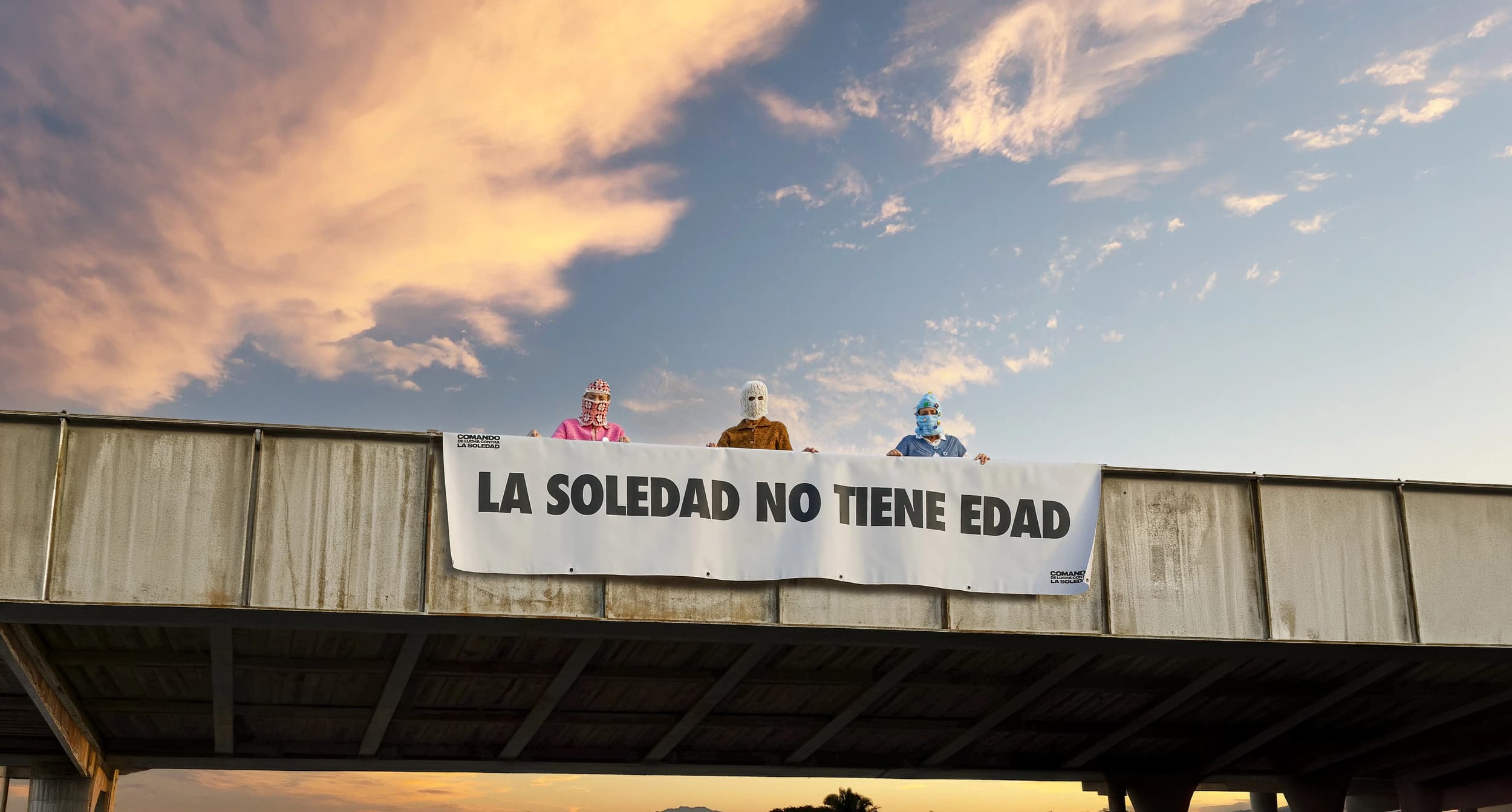 Tres abuelas con pasamontañas en un puente de una autopista con una pancarta que dice "La soledad no tiene edad". La Soledad no tiene edad campaña creativa para la empresa Somos Estupendas. 