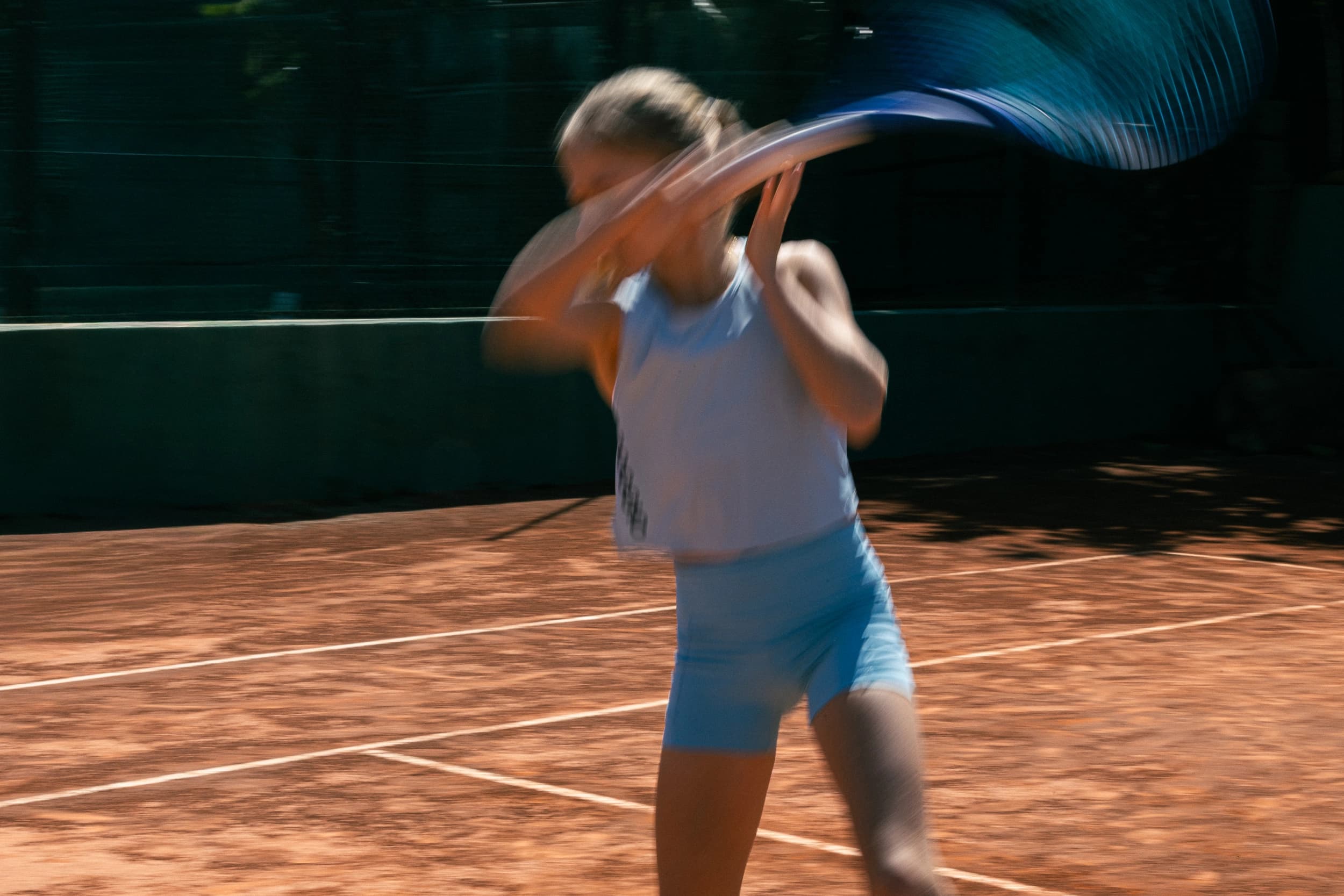 adolescente jugando a tenis en una pista profesional de tierra batida con equipación de la marca ROGGIO de color azul cielo. La imagen está borrosa 