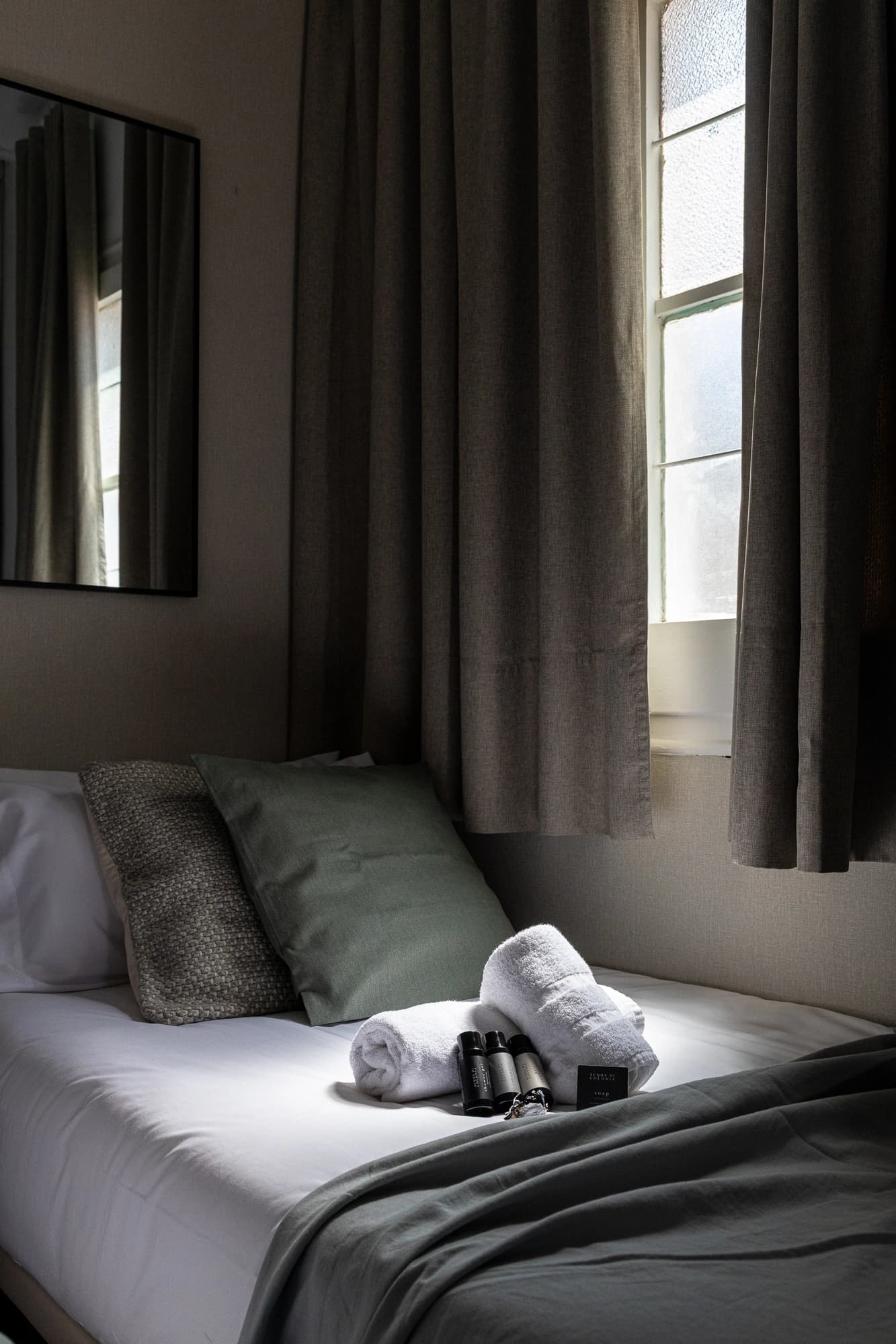 Cama individual con cortinas grises. Luz íntima entra por la ventana de un apartamento de Enville