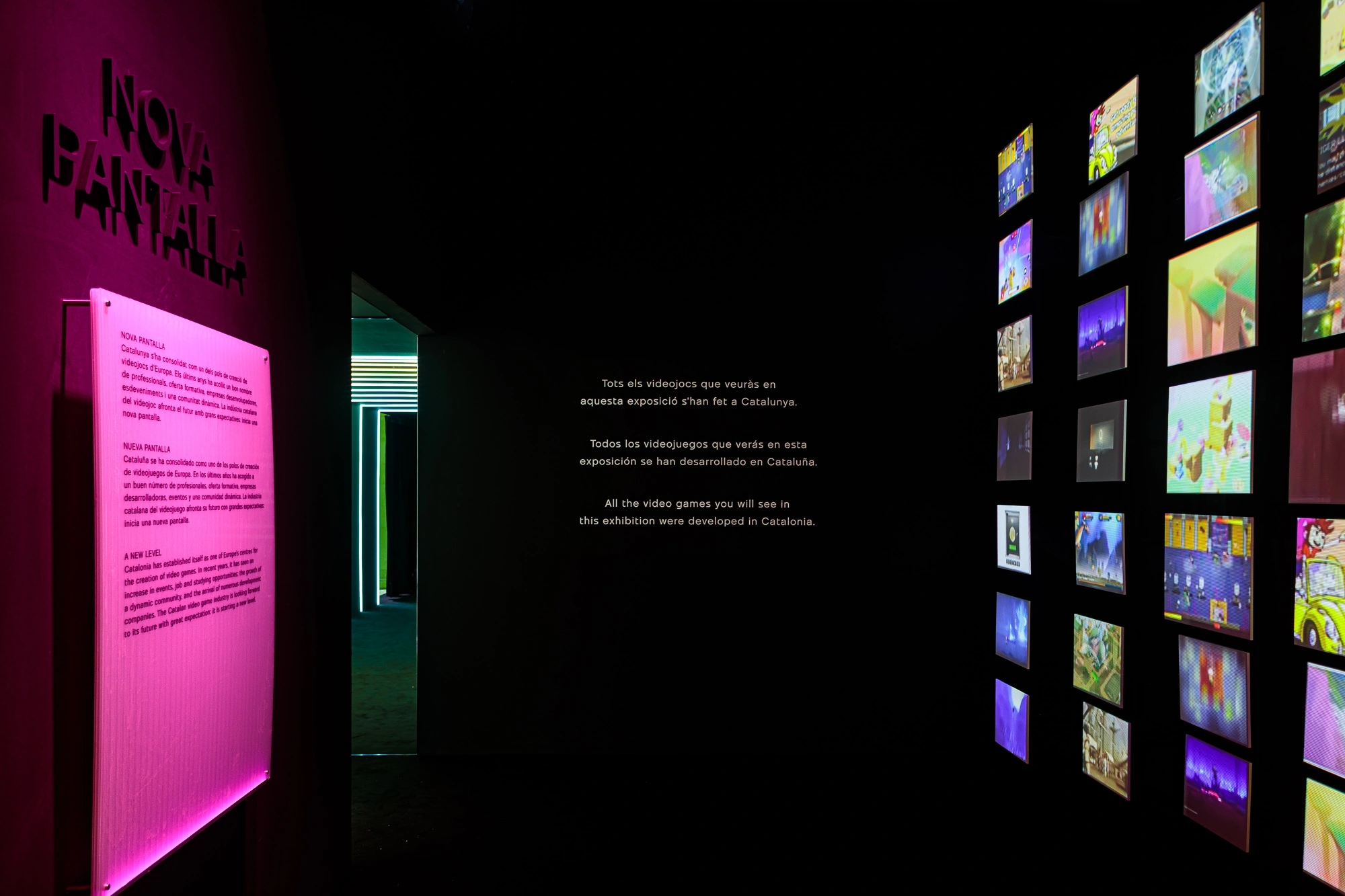 Fotografia de la escenografia de la exposición de Nueva pantalla, el videojuego en Cataluña. Entrada de la exposición, con la cartelería a la izquierda y un montón de pantallitas a la derecha reproduciendo en bucle videojuegos míticos de la indústria.