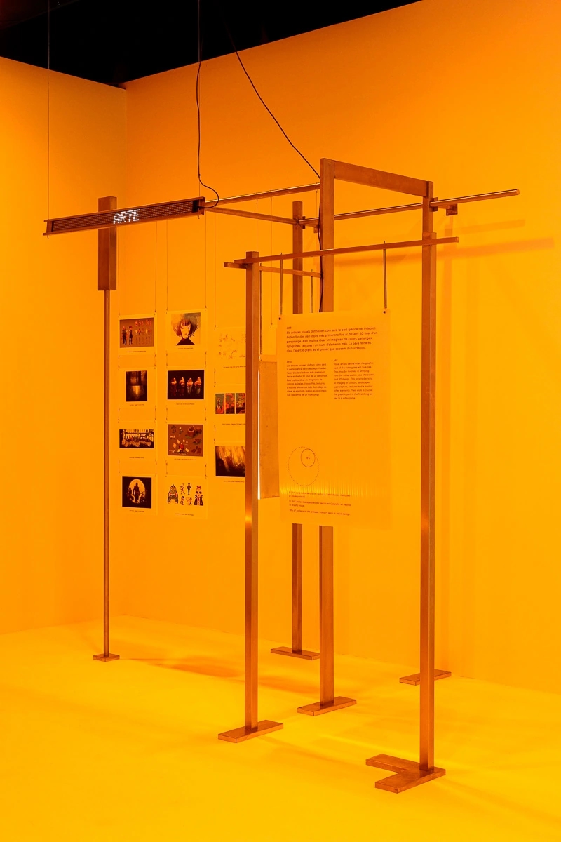 Fotografia de la escenografia de la exposición de Nueva pantalla, el videojuego en Cataluña. Fondo amarillo y muchas actividades tecnológicas.
