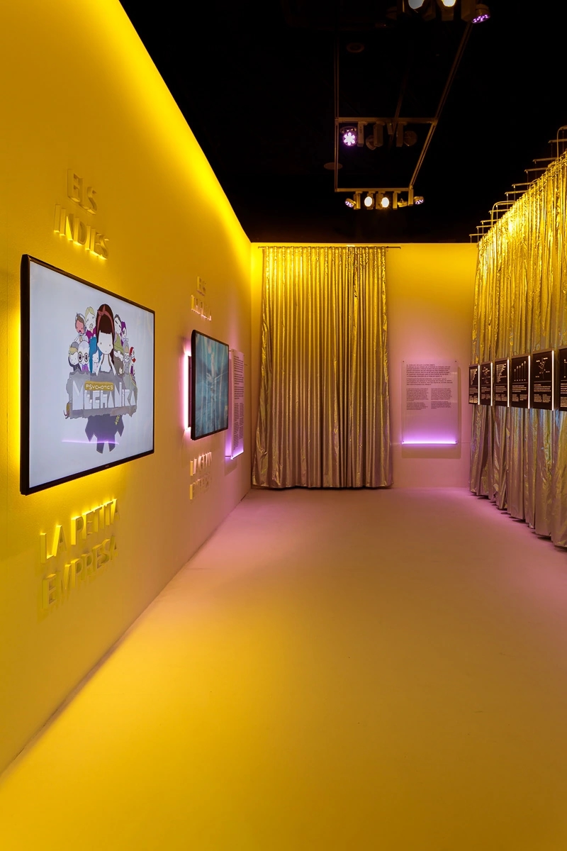 Fotografia de la escenografia de la exposición de Nueva pantalla, el videojuego en Cataluña. Fondo amarillo/rosa y muchas actividades tecnológicas. Hay un televisor a la izquierda reproduciendo vídeos y una cortina plateada de fondo.