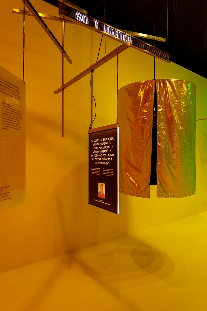 Fotografia de la escenografia de la exposición de Nueva pantalla, el videojuego en Cataluña. Fondo amarillo/verde y una cápsula de aislamiento acústico para escuchar sonidos relacionados con el videojuego.
