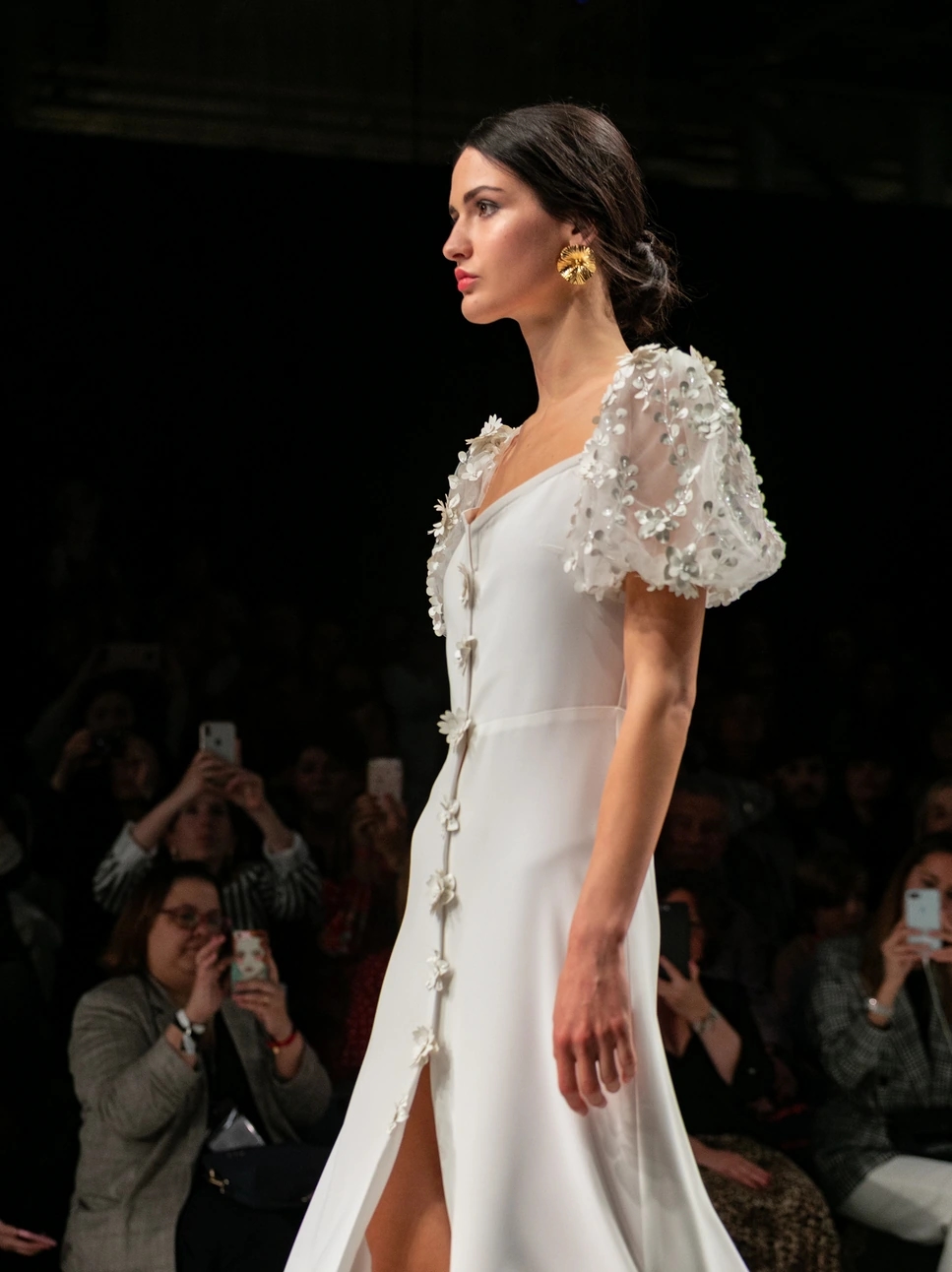 Modelo con vestido fino de novia andando de perfil con un moño bajo y unos pendientes de oro para el desfile de Beba's en la Barcelona Bridal Fashion Week.