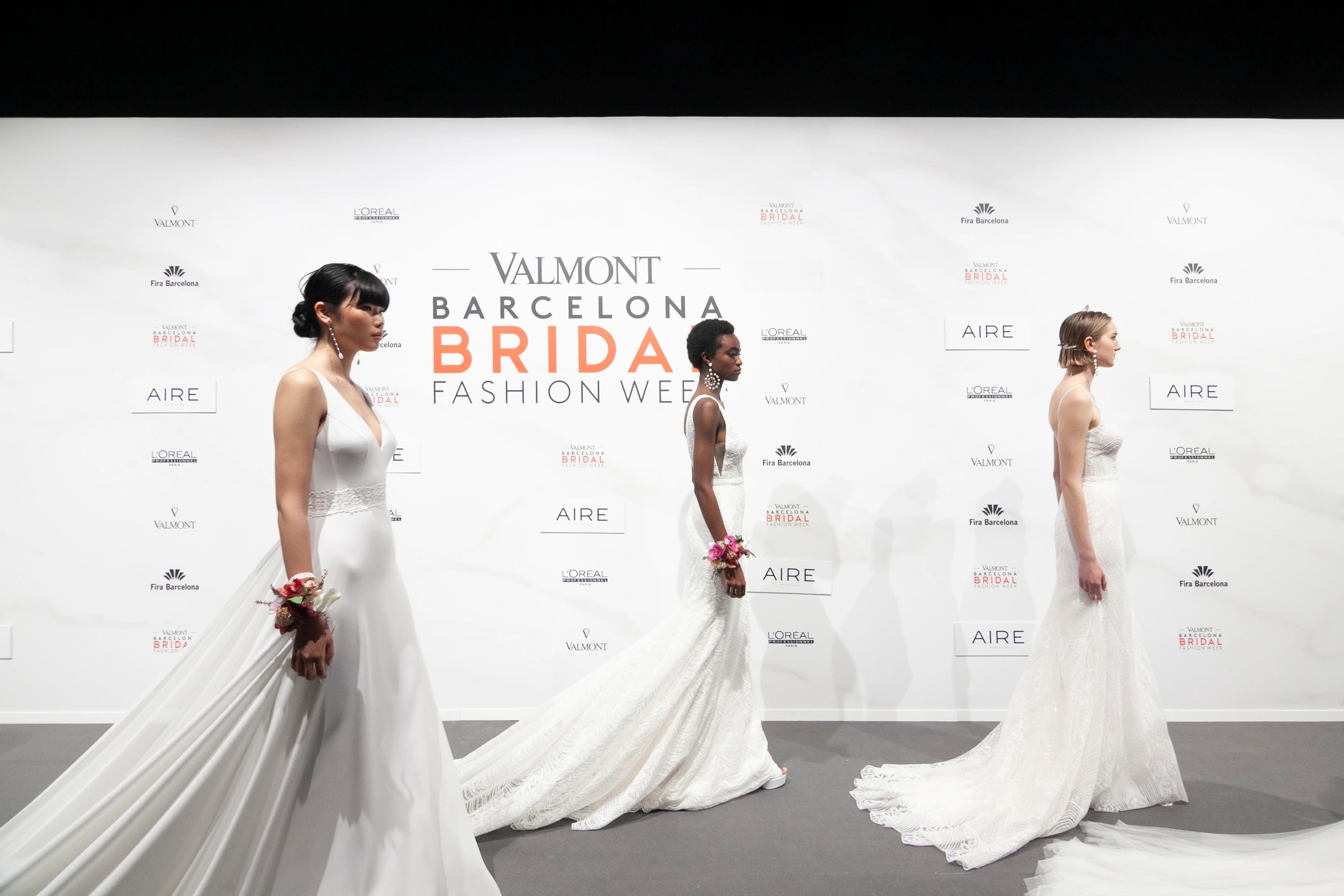 3 modelos esperando a entrar en el desfile de AIRE, con el cartel de Valmont Barcelona Bridal Fashion Week detrás.