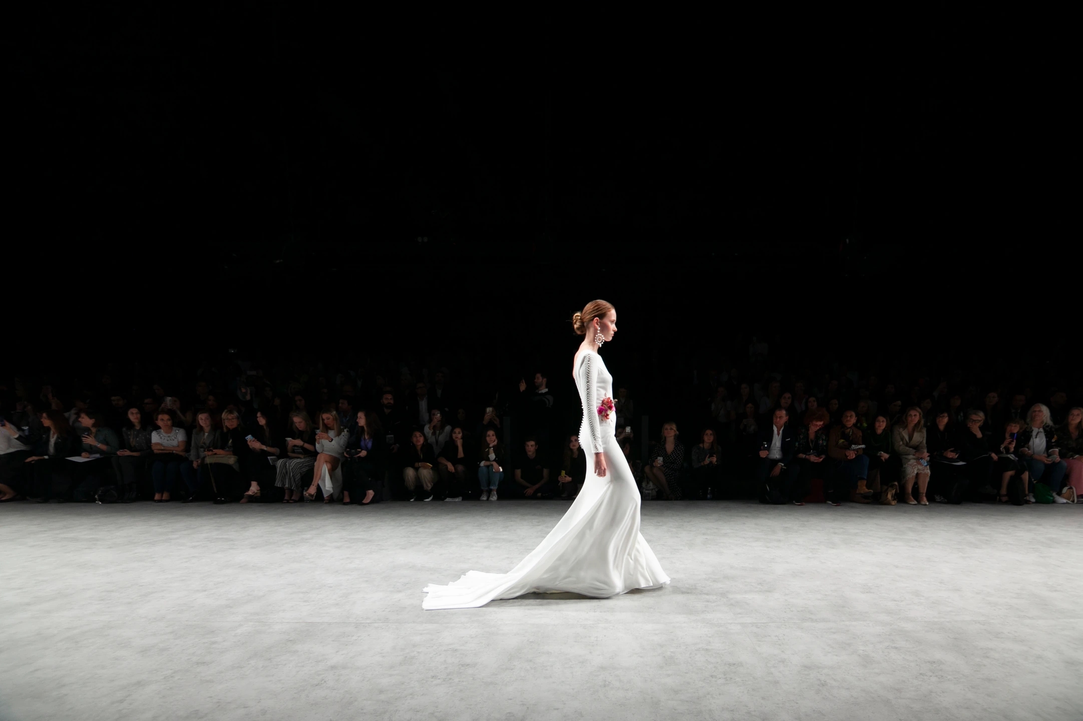 Modelo andando de perfil en el desfile de novias de AIRE Barcelona, se ven de fondo los visitantes sentados. Pasarela de Valmont Barcelona Bridal Fashion Week.