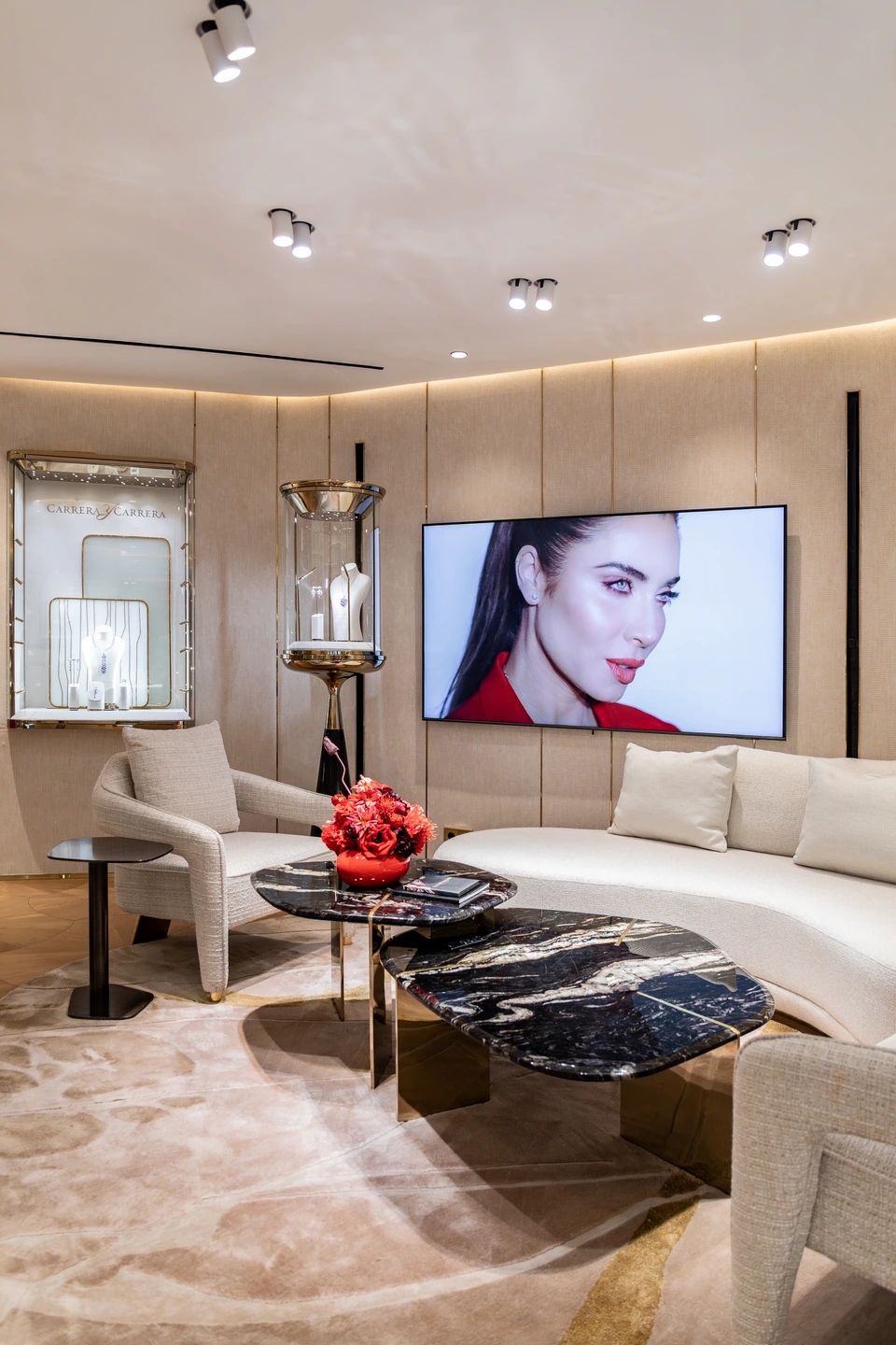 Espacio de la planta superior donde se observa un sofá confortable de color crema un sillón del mismo color un centro de mesa con flores rojas y la mesa en marmolado negro, con una pantalla apoyada en pared donde se ve a Pilar Rubio donde muestra la colección de la joyería