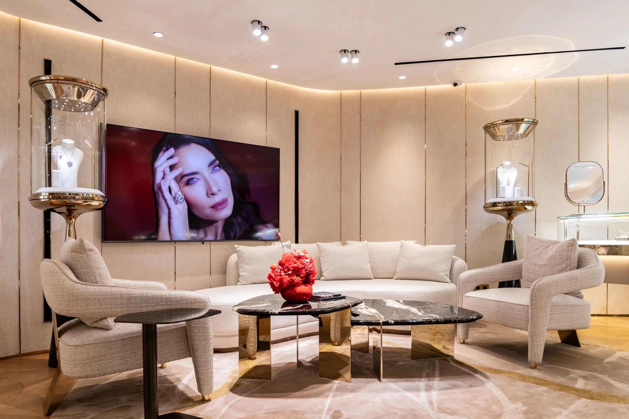 Espacio de la planta superior donde se observa un sofá confortable de color crema un sillón del mismo color un centro de mesa con flores rojas y la mesa en marmolado negro, con una pantalla apoyada en pared donde se ve a Pilar Rubio donde muestra la colección de la joyería