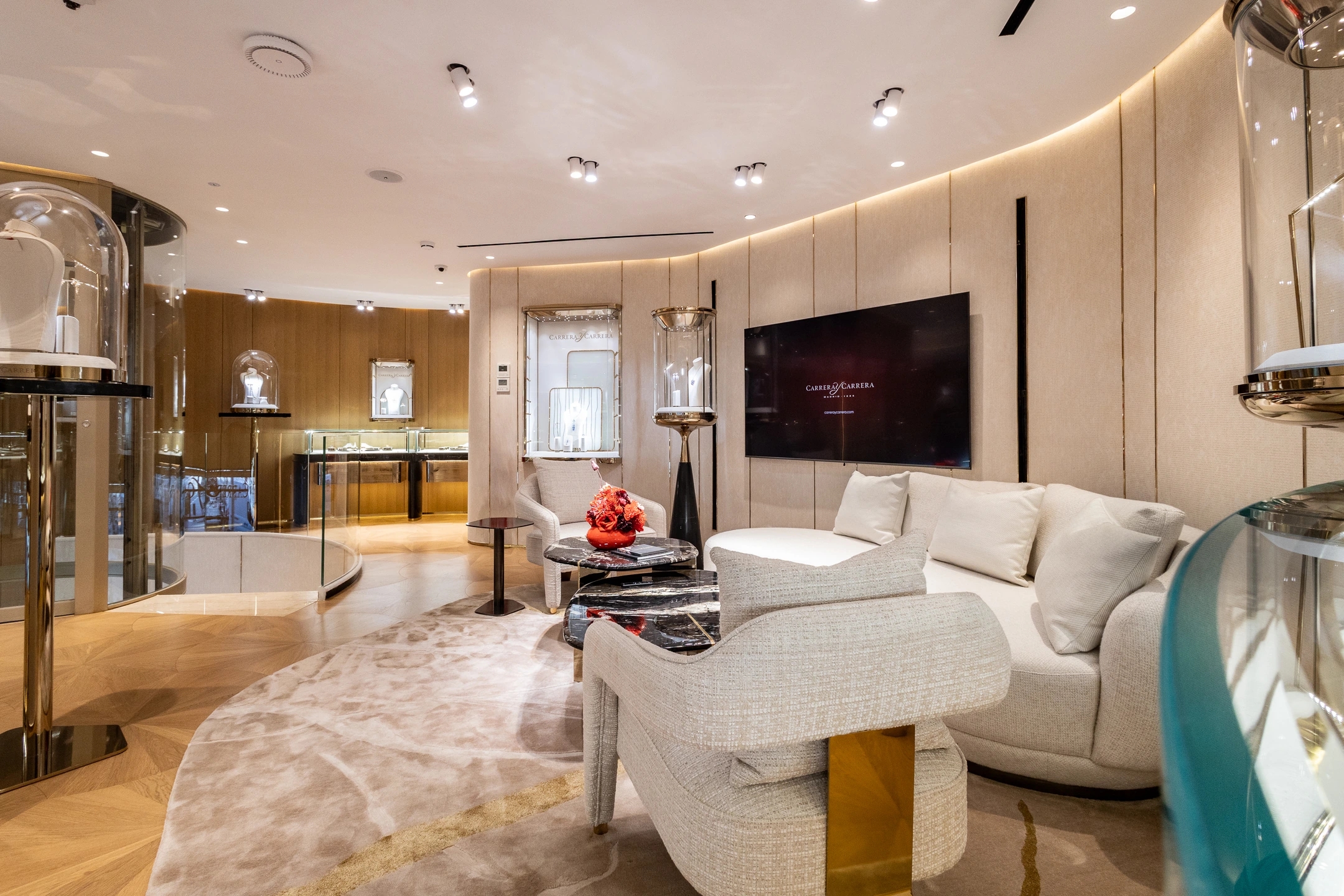Espacio de la planta superior donde se observa un sofá confortable de color crema un sillón del mismo color un centro de mesa con flores rojas y la mesa en marmolado negro, con una pantalla apoyada en pared donde se ve a Pilar Rubio donde muestra la colección de la joyería