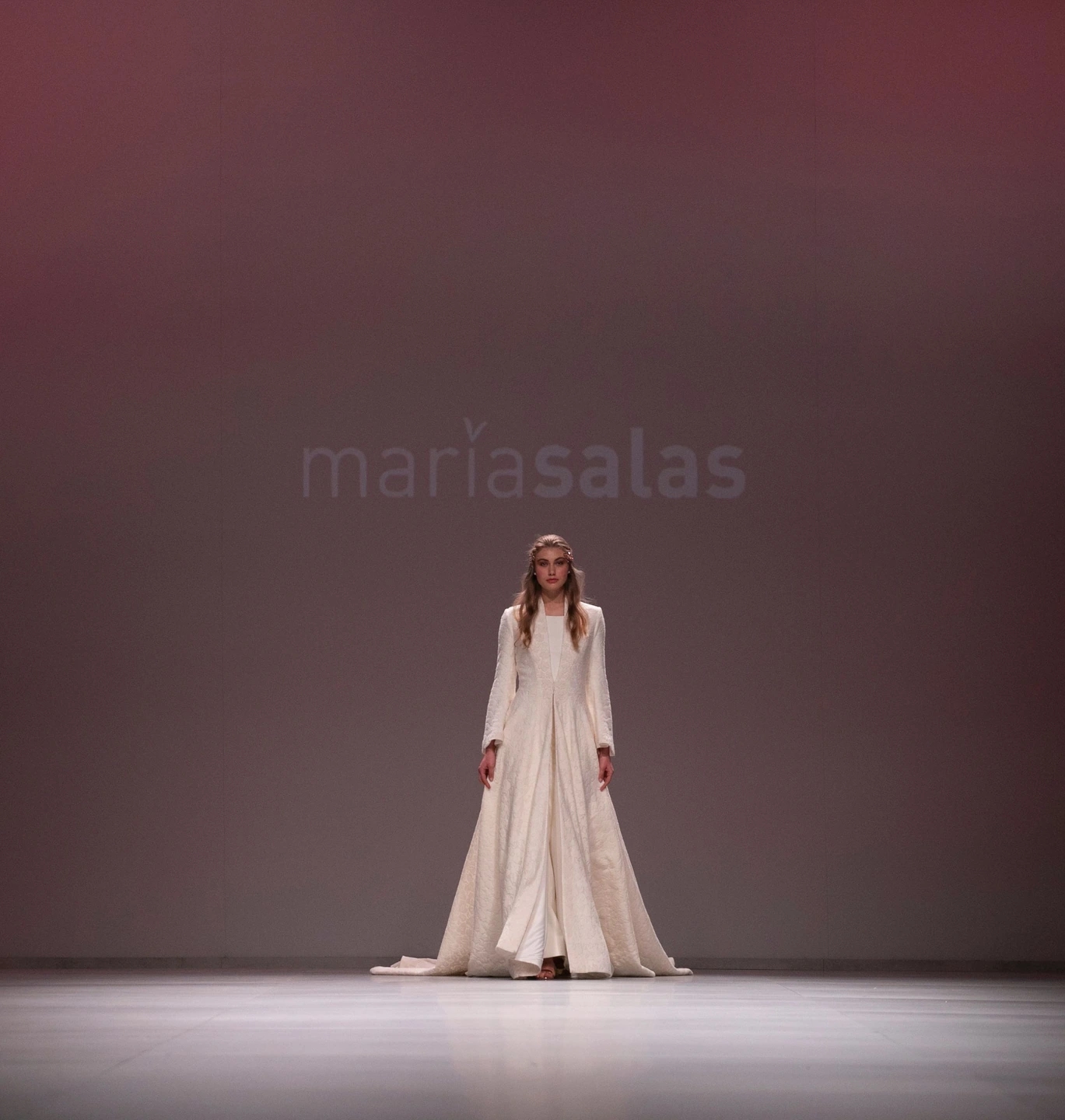 Modelo con vestido de novia en pose de elfa andando de frente con capa en el desfile de María Salas para la Barcelona Bridal Fashion Week.
