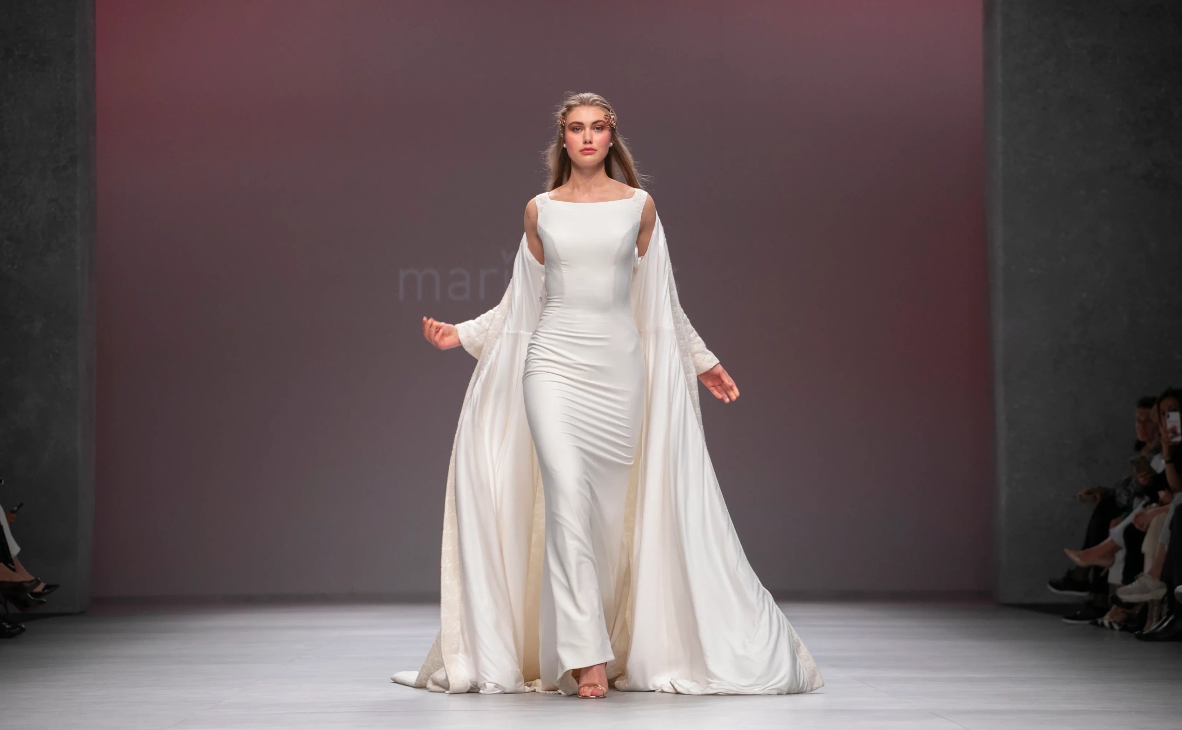 Modelo con vestido de novia en pose de diosa andando de frente con capa en el desfile de María Salas para la Barcelona Bridal Fashion Week.